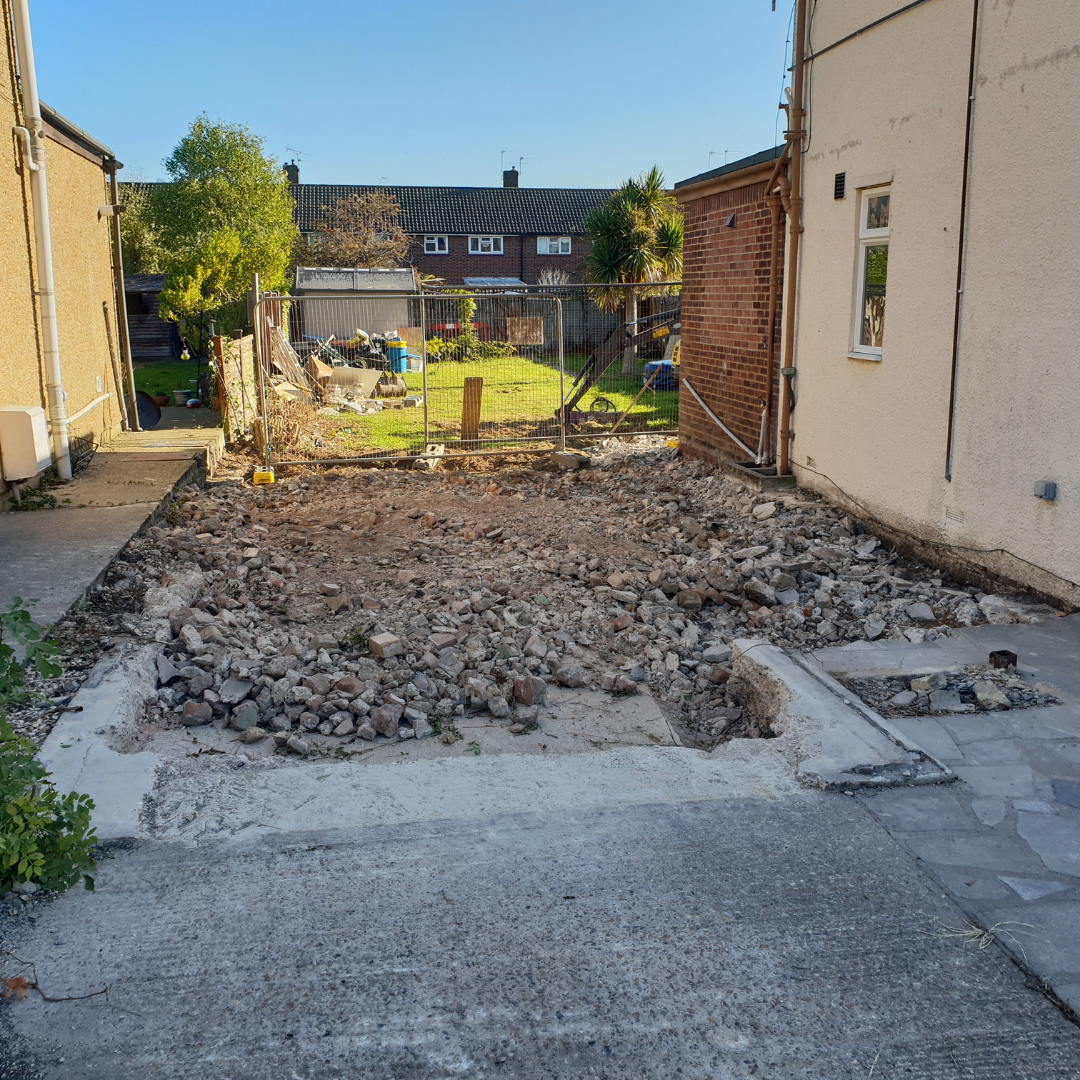 A construction site between two residential buildings showing a patch of rubble and soil cleared for new work.