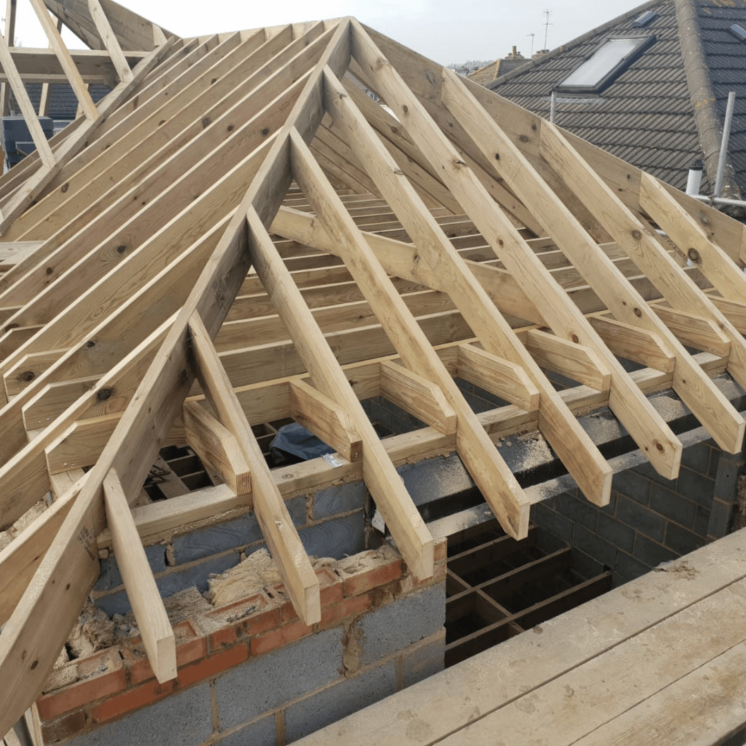 Wooden roof framing in progress, featuring rafters and a hip beam extending from a blockwork wall on a construction site.