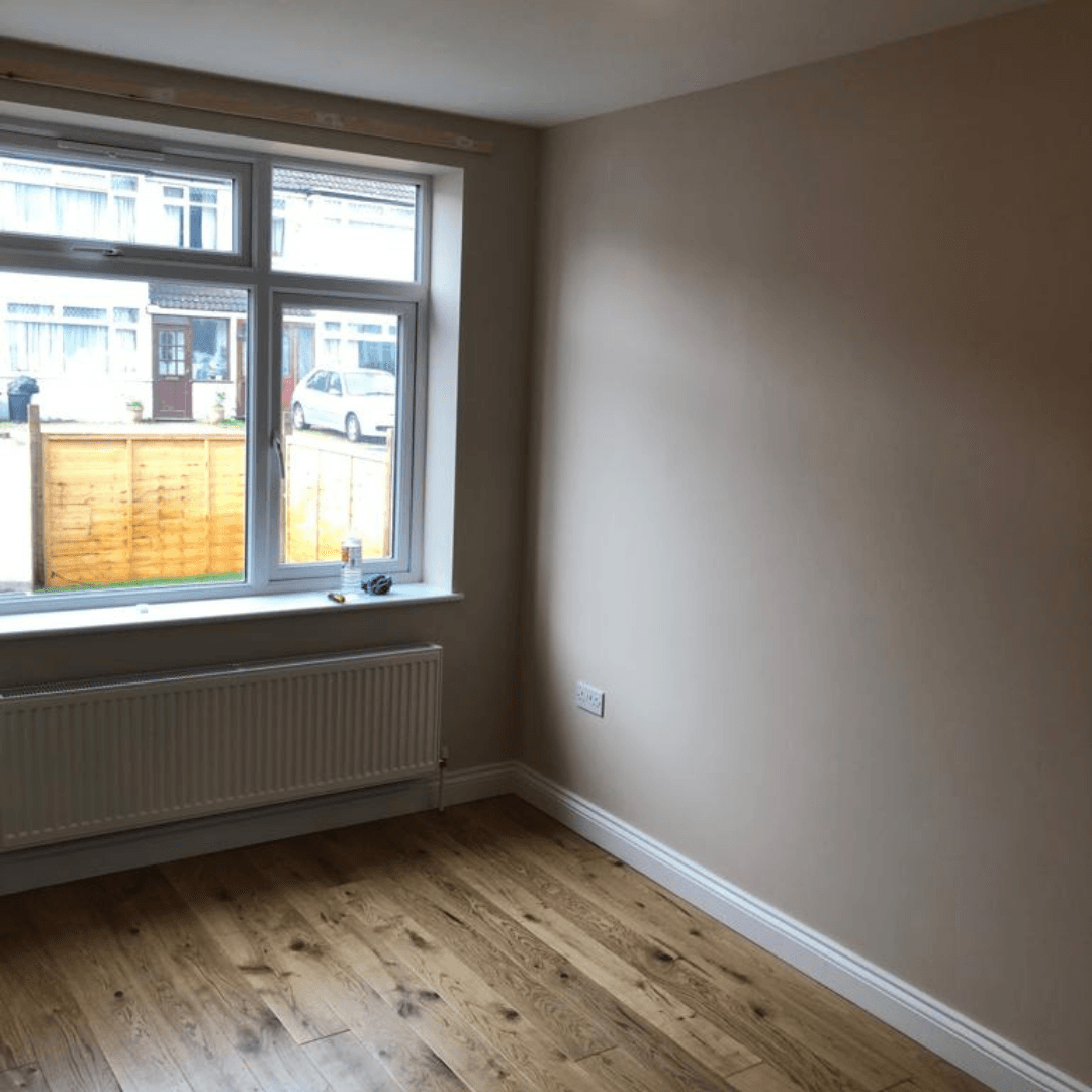 An empty, sunlit room with beige walls, a window overlooking a street, a white radiator, and wood-grain flooring.