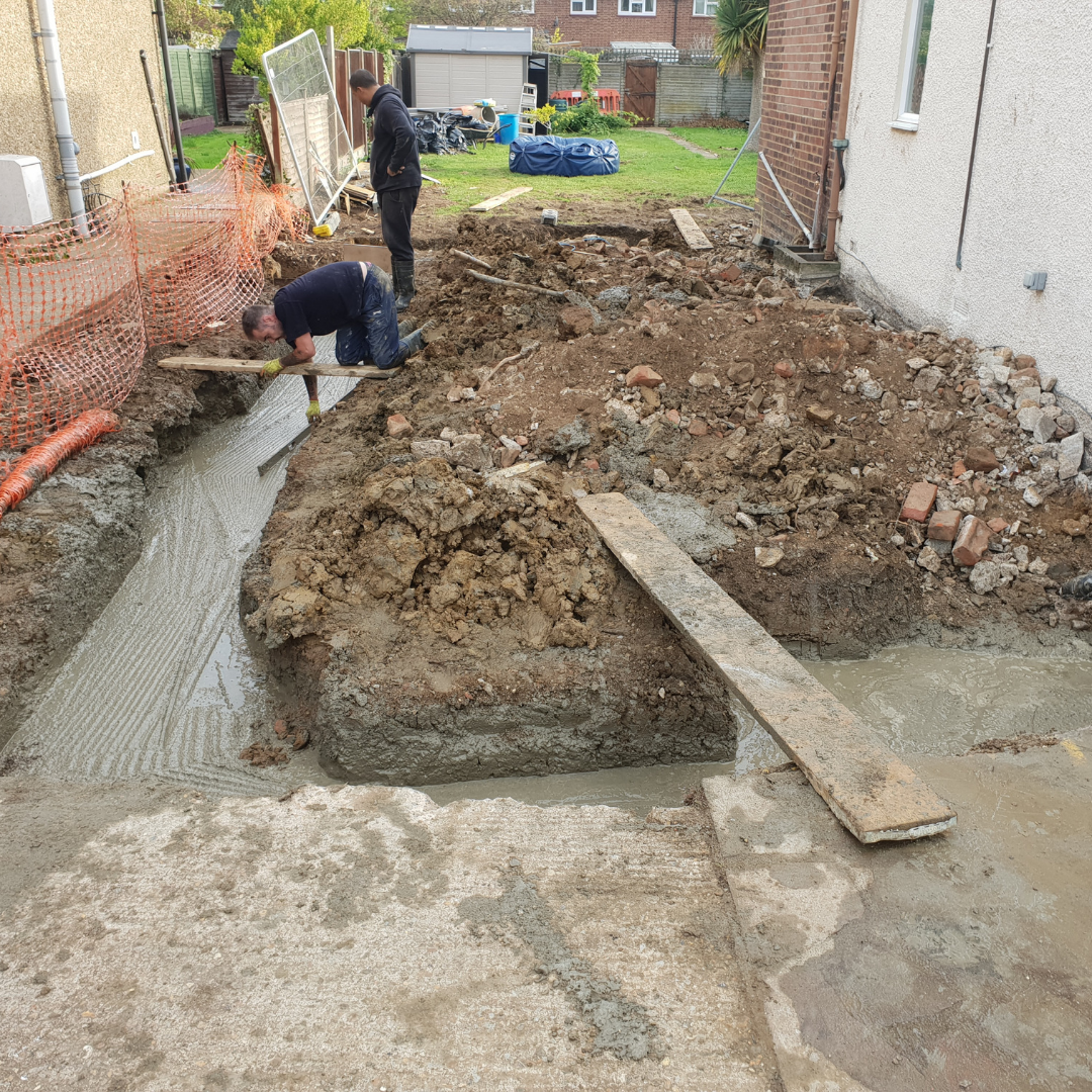 Two workers prepare a foundation trench with wet concrete alongside a house exterior during a construction project.