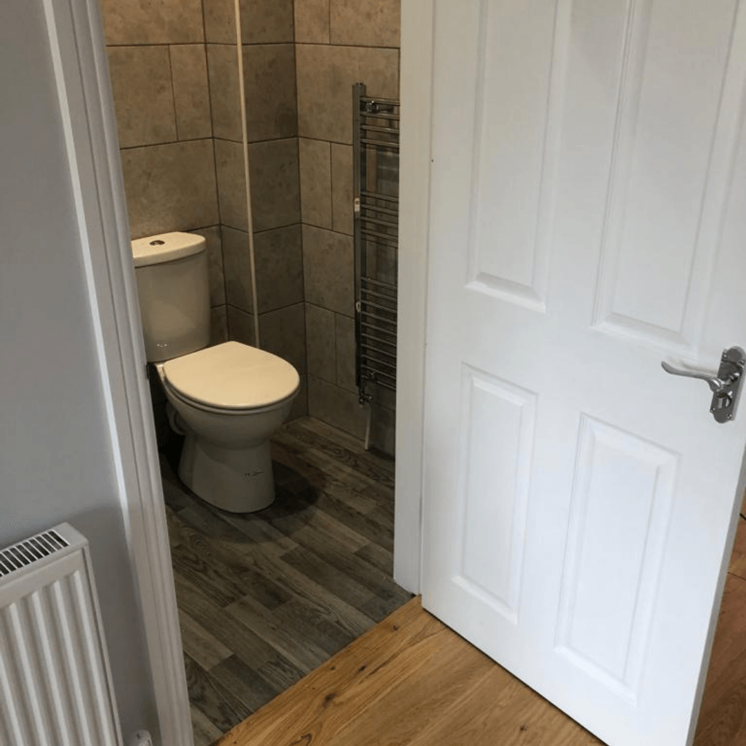 A toilet in a small alcove with grey tiled walls, dark wood-look flooring, and a white door in the foreground.