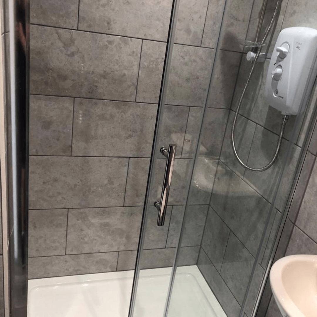 A walk-in shower with grey rectangular tiles, a glass door, and a white electric shower unit mounted on the wall.