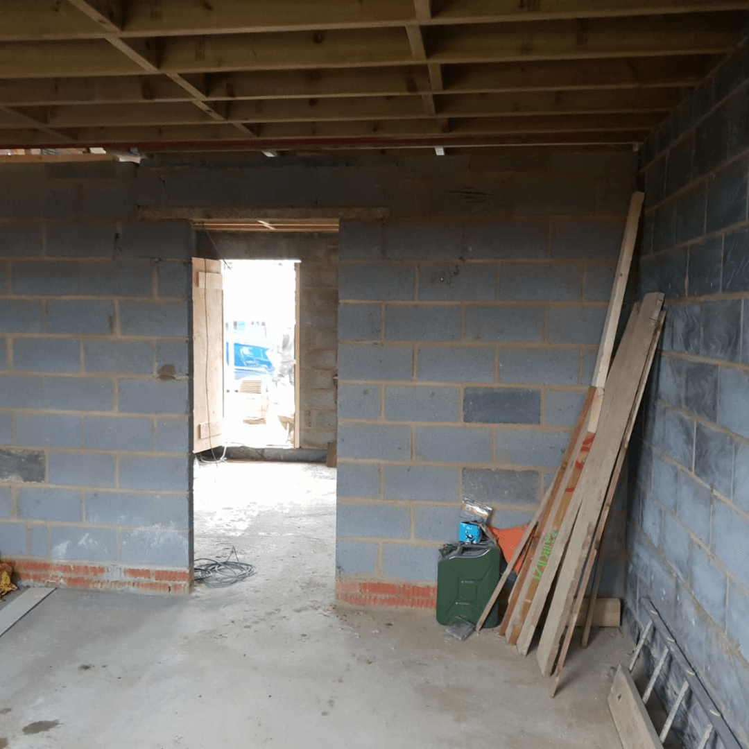 An interior construction site showing a doorway leading to daylight, grey block walls, and wooden joists above.
