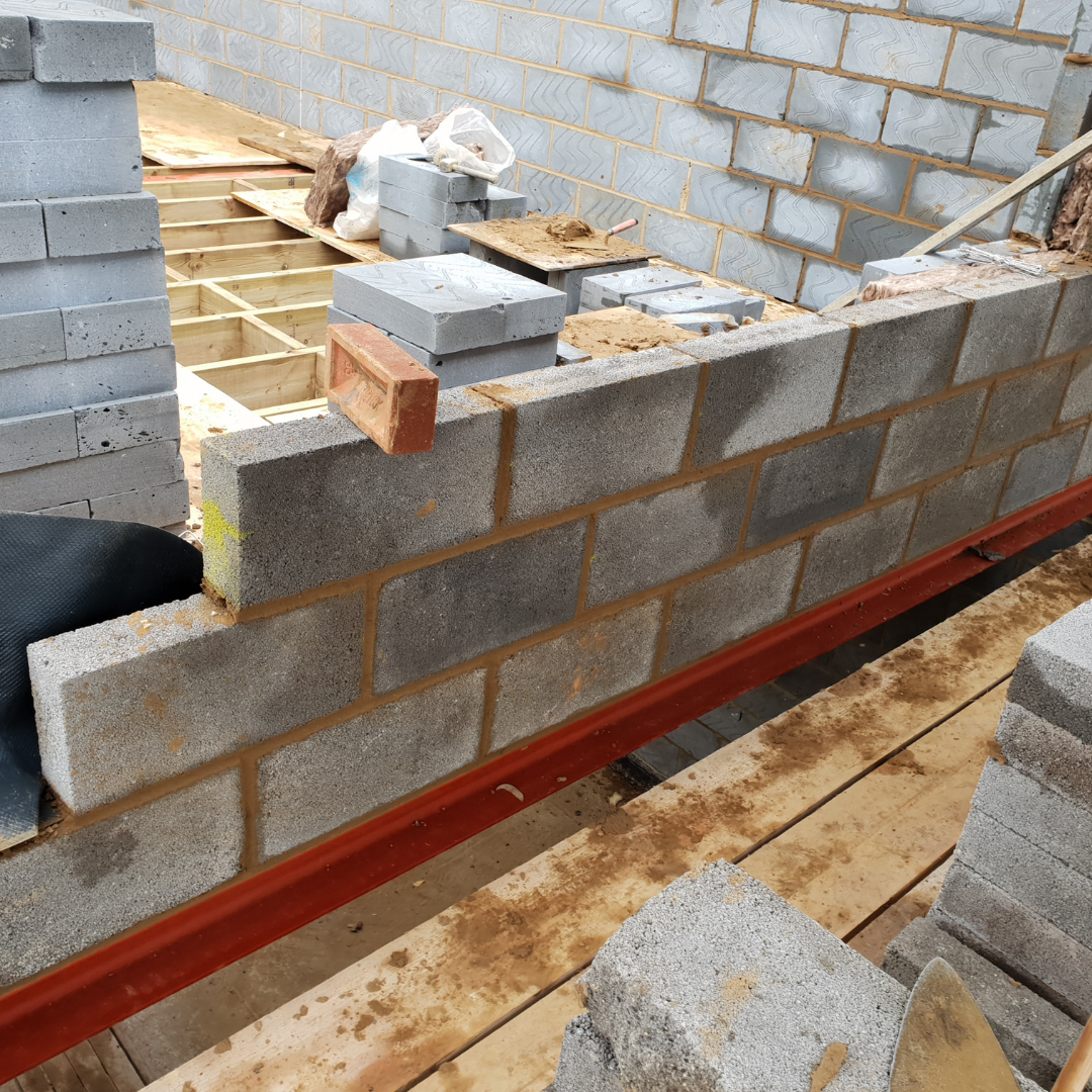 A construction site featuring a partially built concrete block wall resting on a red steel beam.