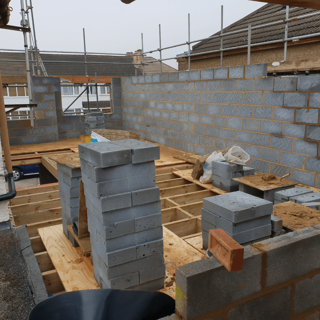 An outdoor construction site showing a wooden floor frame, concrete block walls, and stacks of blocks on scaffolding.