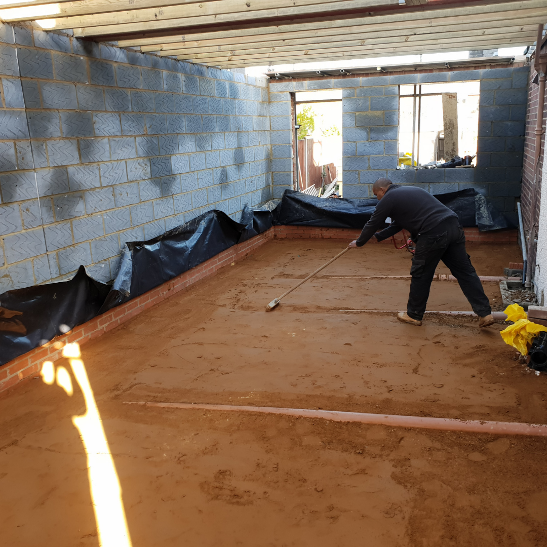 A person uses a long-handled tool to level reddish sand inside an unfinished room with grey block walls and black sheeting.