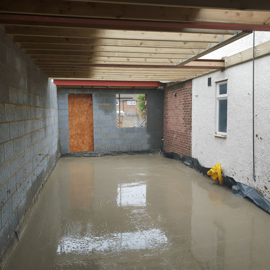 An unfinished room addition with a wet concrete floor, wooden roof joists, and exposed brick and block walls.