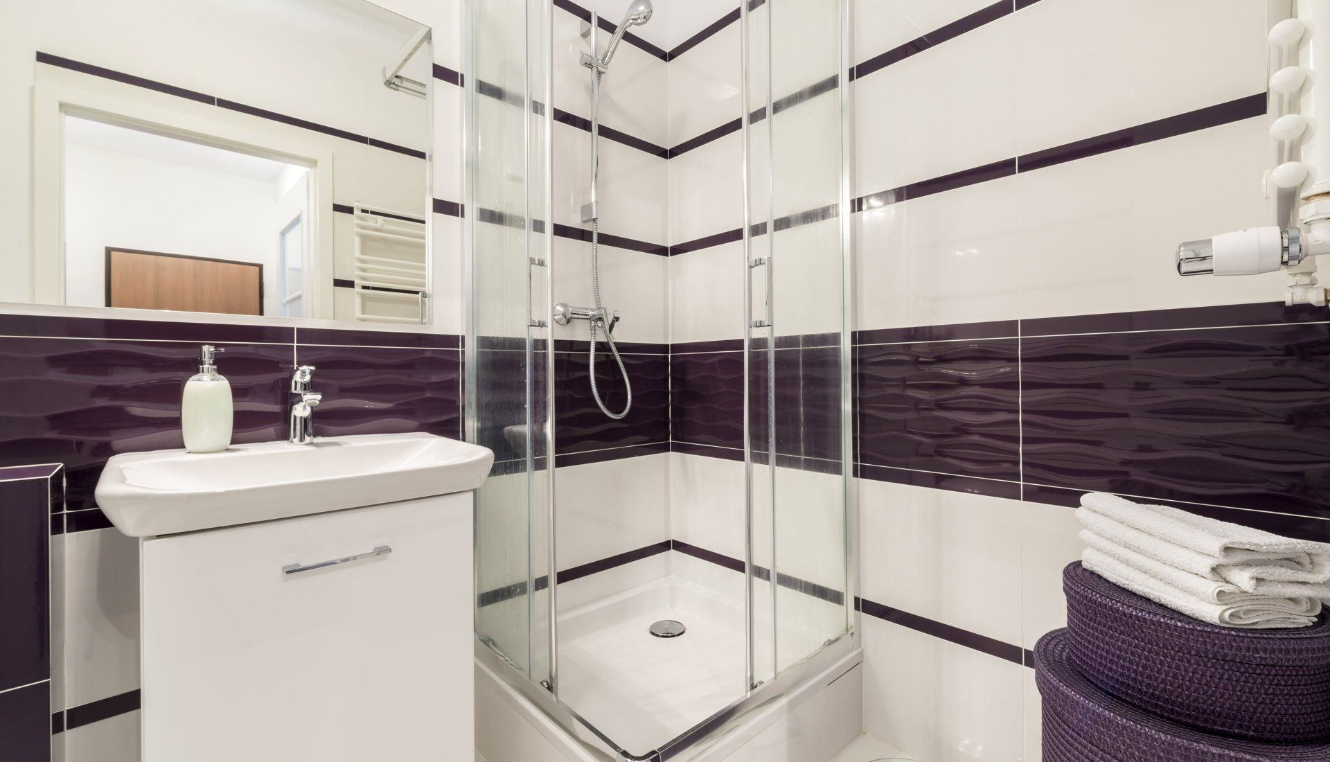 A modern bathroom with a glass shower, white sink, and white-and-purple horizontal striped tiled walls.