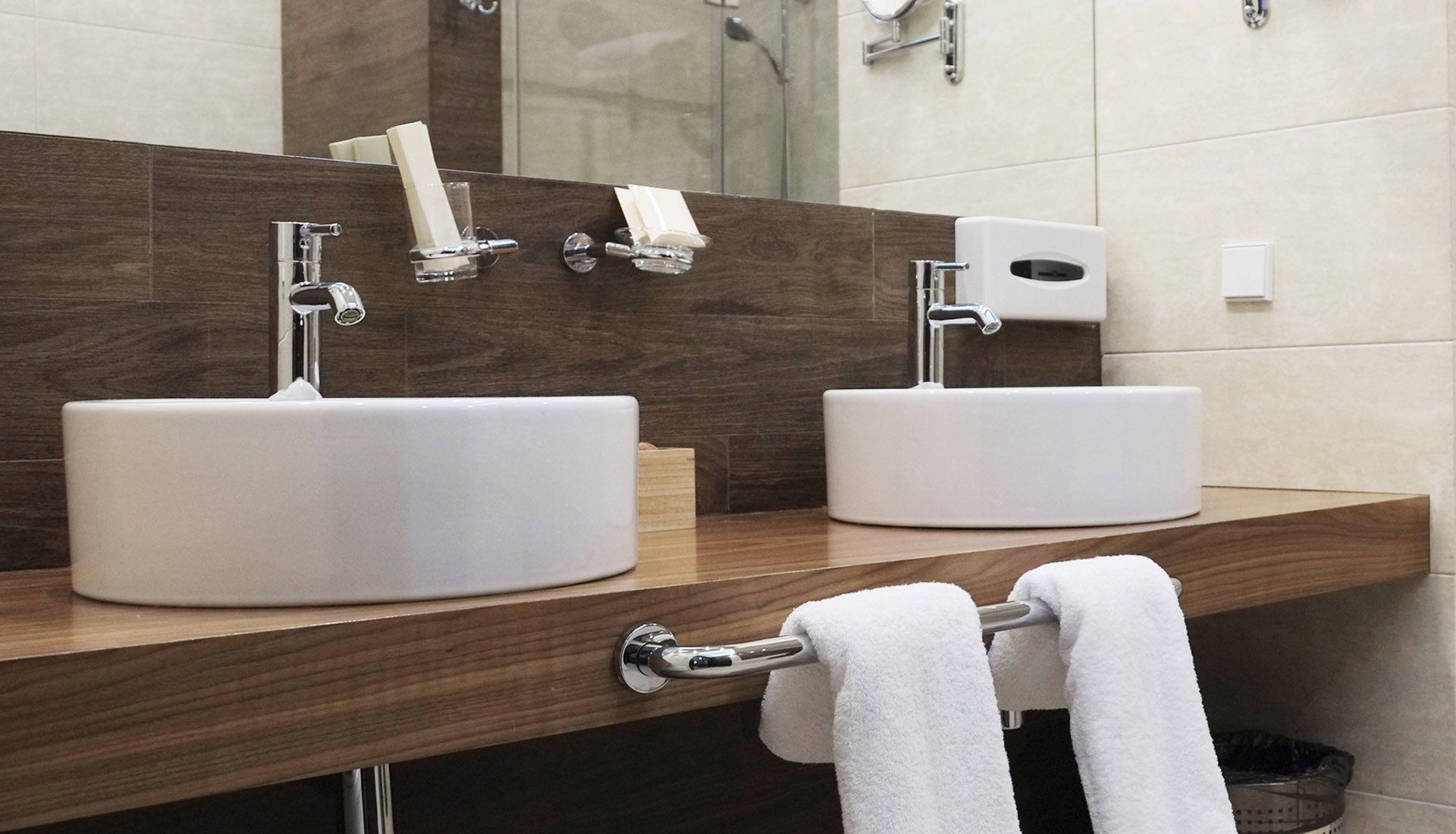 A modern hotel bathroom vanity with two white basin sinks, chrome faucets, and white towels hanging on a wooden counter.