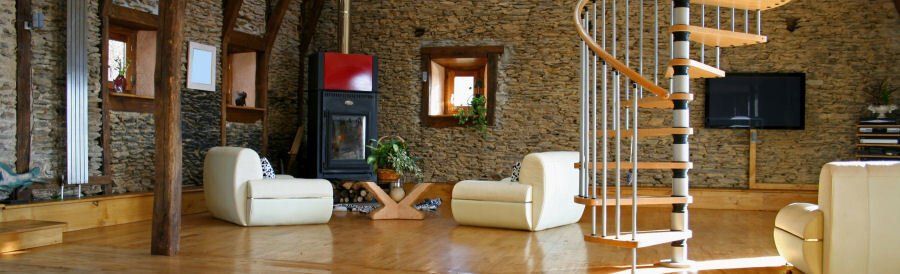 Hillrose Property Service Homepage Image A spacious room with stone walls, wooden flooring, white armchairs, a modern fireplace, and a spiral staircase.