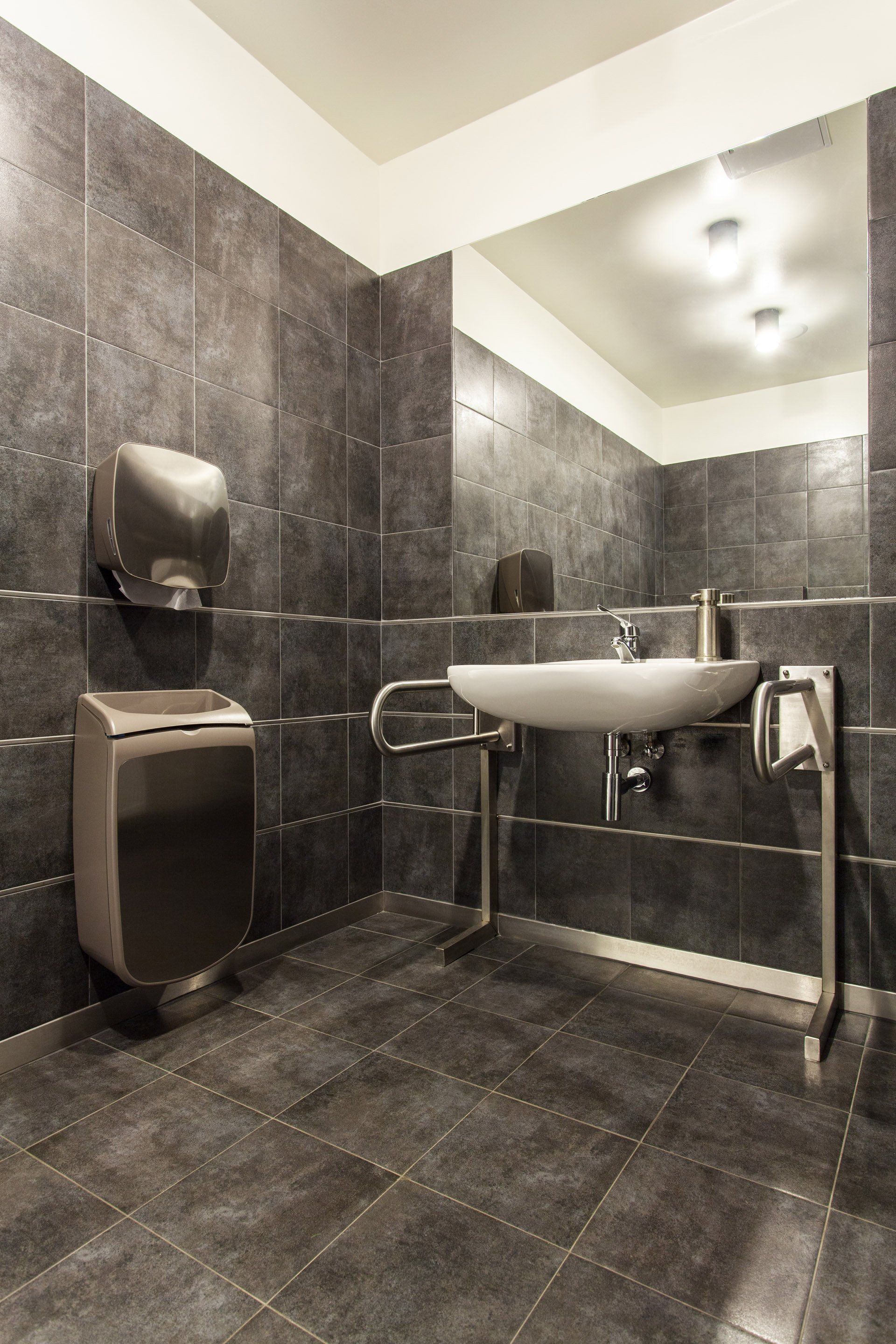 Accessible restroom with a wall-mounted sink, support handrails, and a hand dryer against dark gray tiled walls.