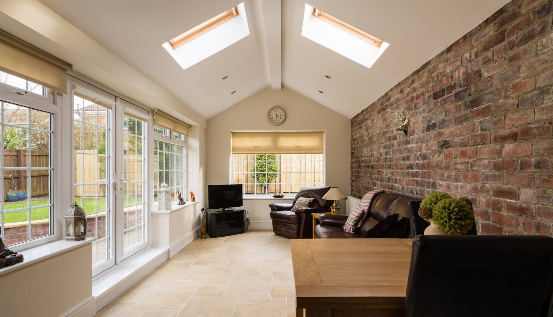 A sunroom with a vaulted ceiling, skylights, exposed brick wall, tile flooring, leather furniture, and large windows.