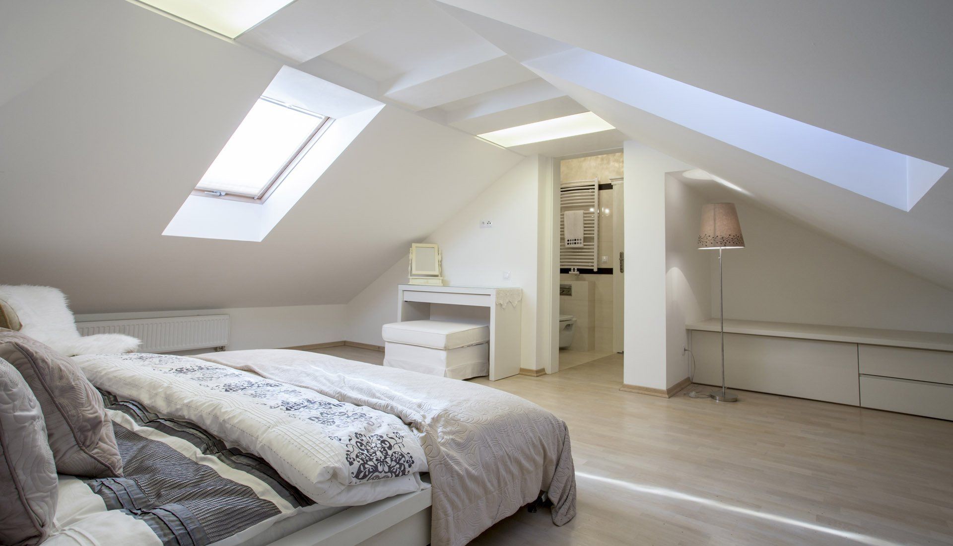 A bright attic bedroom with white slanted ceilings, a skylight, a made bed, a small desk, and a doorway to a bathroom.