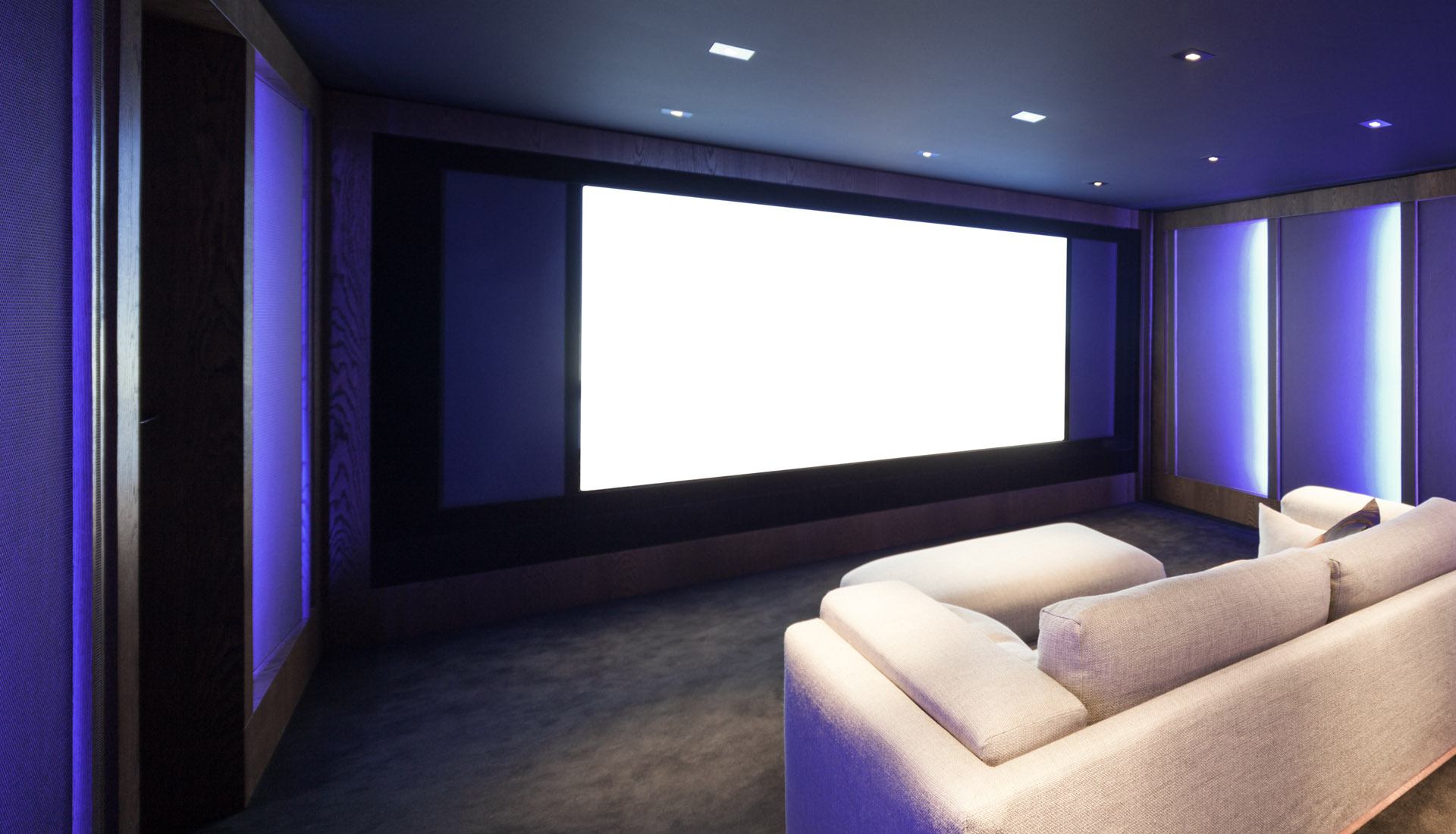 A modern home theater with a large white screen, dim blue mood lighting, and comfortable white sofas on a dark floor.