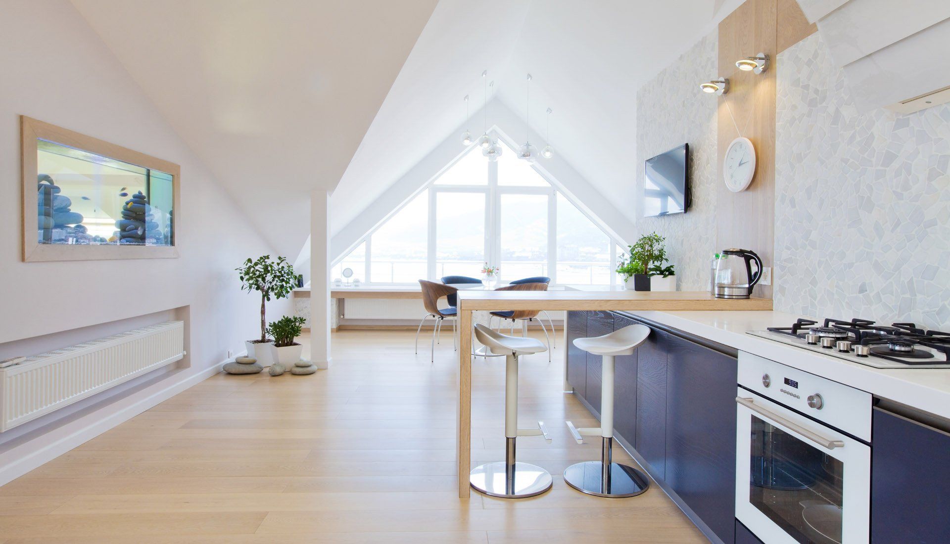 Modern open-plan kitchen and dining area in an attic with light wood floors, blue cabinets, and a tall gable window.