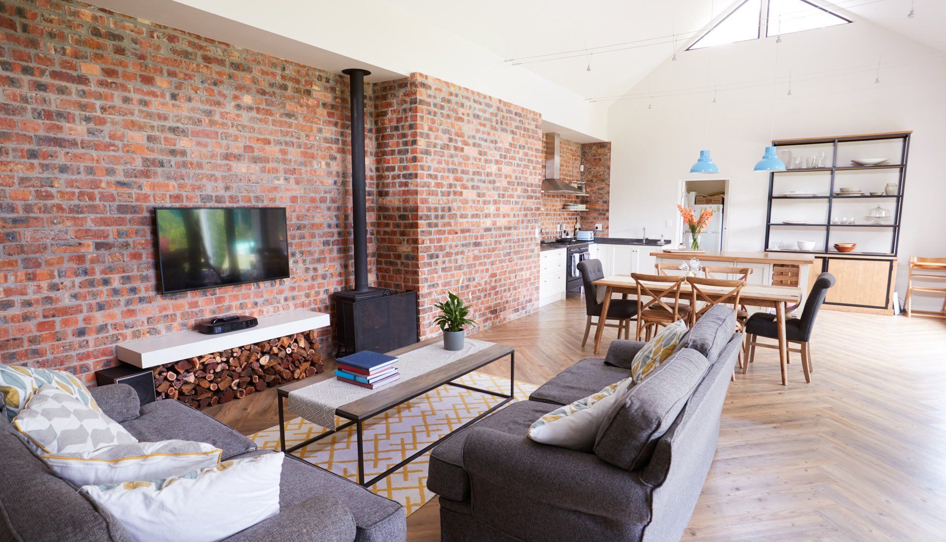 A living room with an exposed brick wall, a TV, a wood-burning stove, gray sofas, and a dining area in an open floor plan.