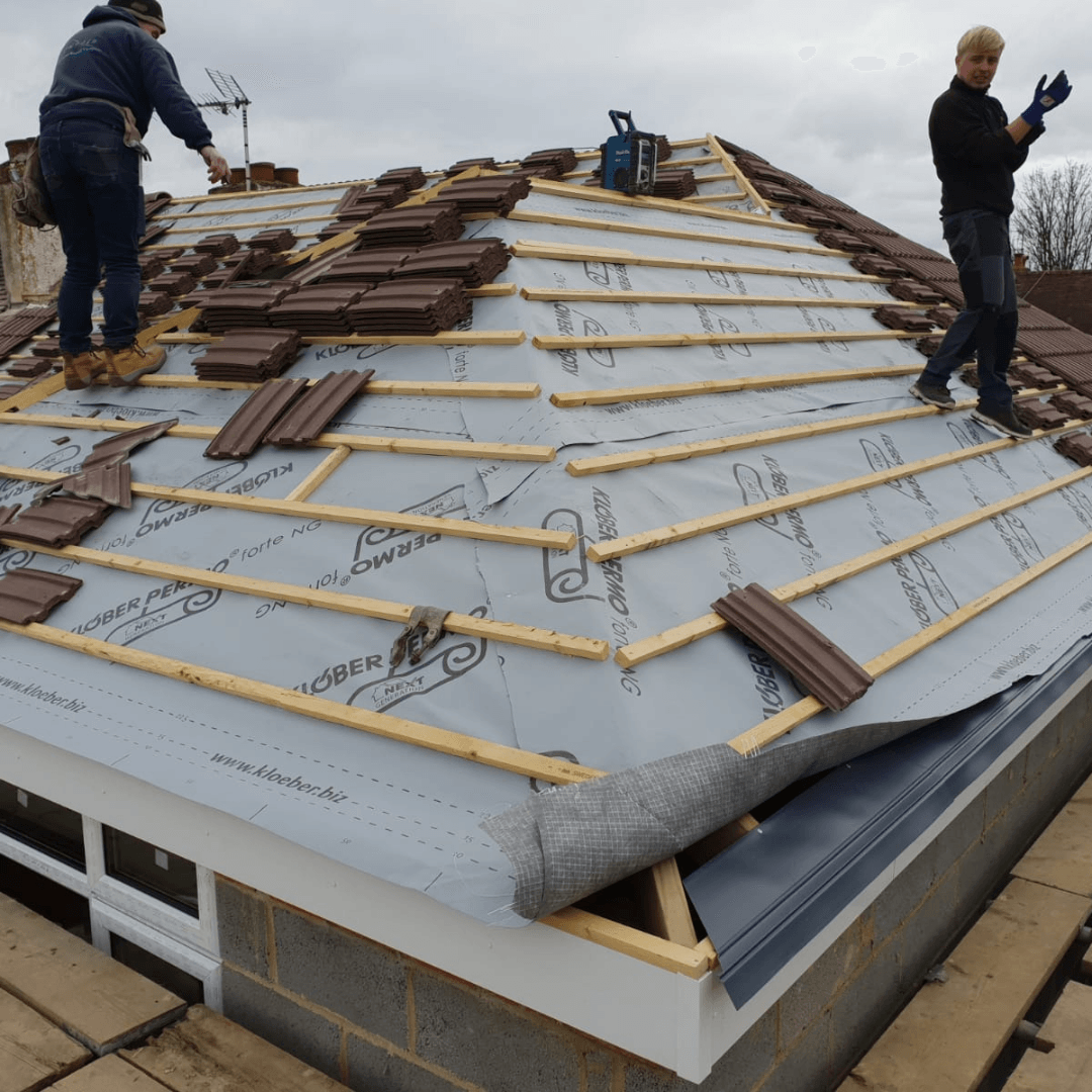 Two people work on a residential roof, installing brown clay tiles over wooden battens and a grey moisture barrier.