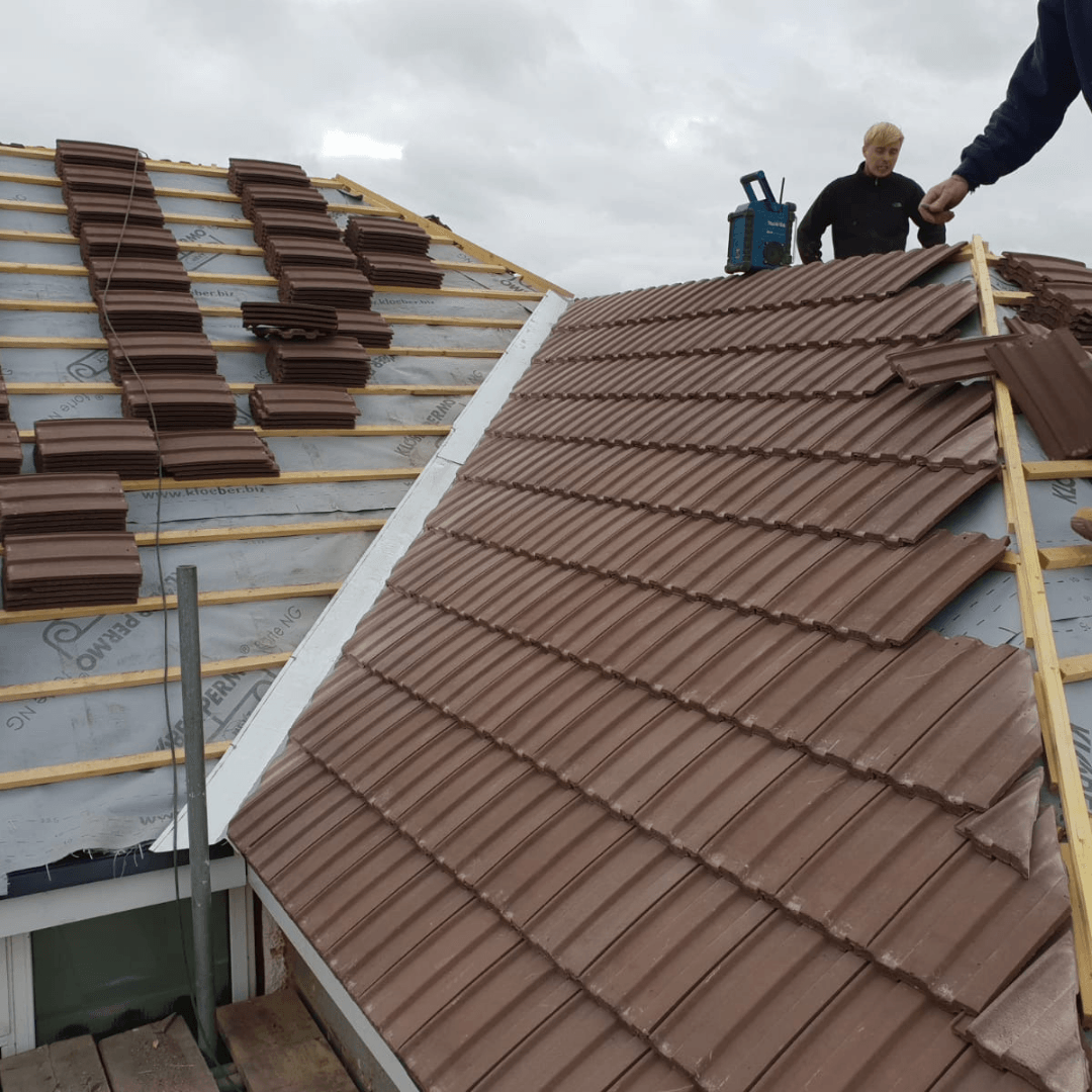 A person working on a roof, installing brown tiles on a wooden frame over a light-colored protective underlay.