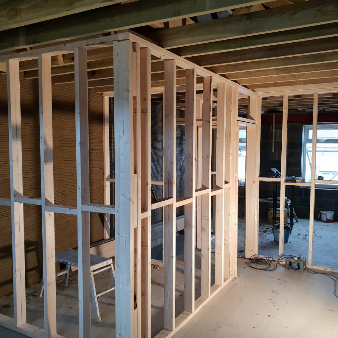 Wooden wall framing structure in an interior construction site with exposed floor joists and a window in the background.