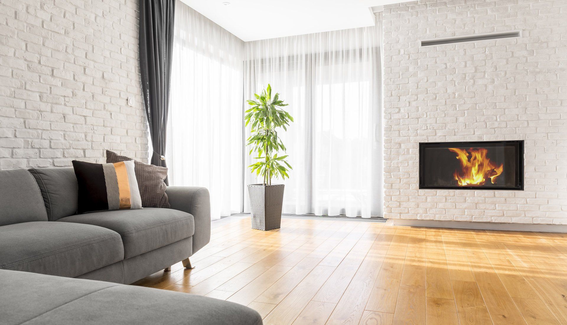 A bright living room with a grey sofa, light wood floors, a potted green plant, and a fireplace in a white brick wall.