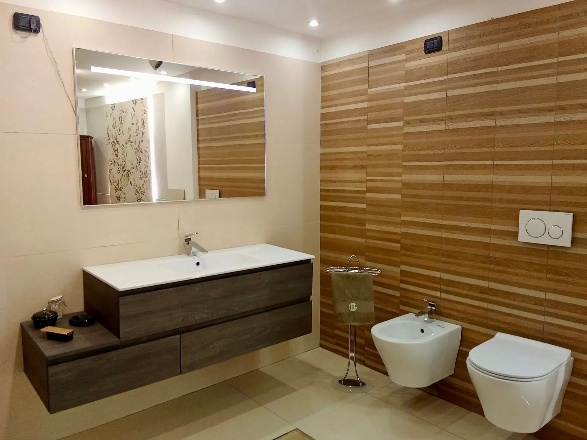 Bagno moderno color beige con mobile lavabo in legno, specchio, WC sospeso, bidet e box doccia in vetro.