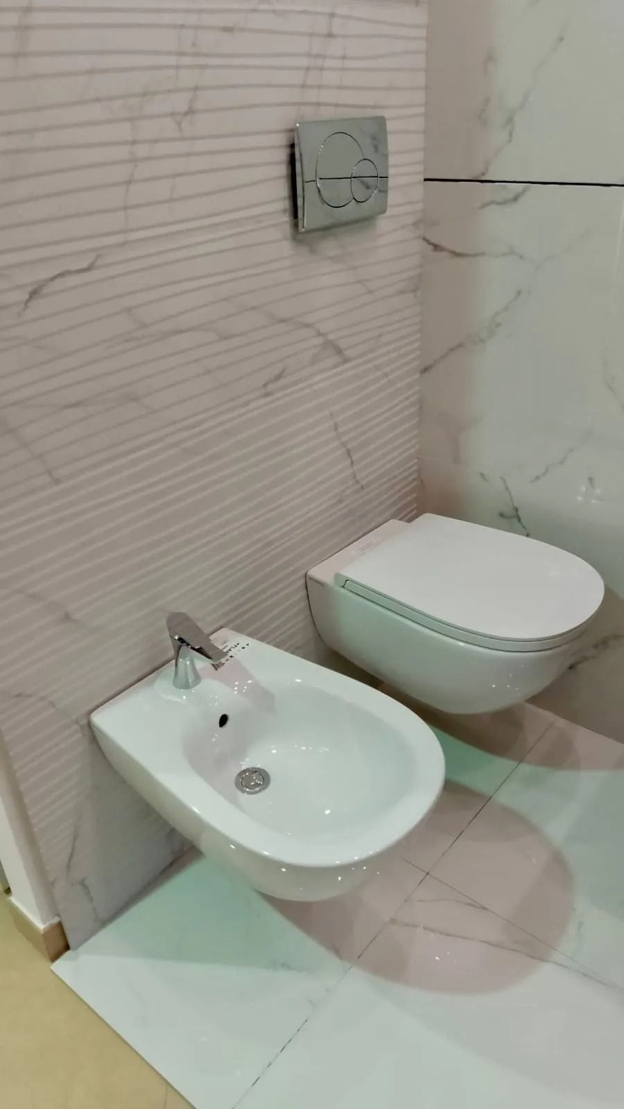 Piccolo bagno moderno con WC sospeso, bidet bianco e pareti rivestite con piastrelle beige.