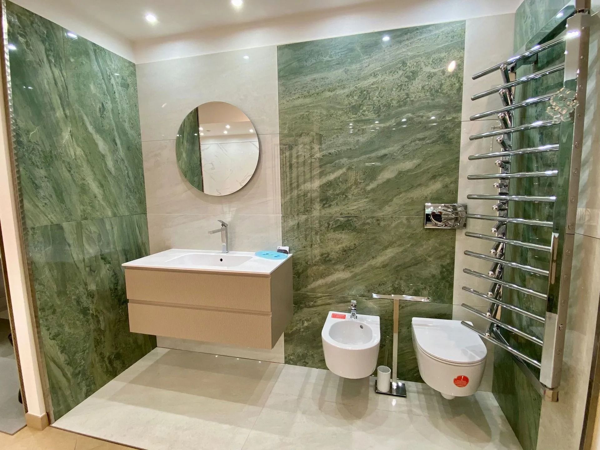 Bagno moderno con pareti in marmo verde, mobile lavabo sospeso in legno, specchio rotondo, WC, bidet e scaldasalviette.