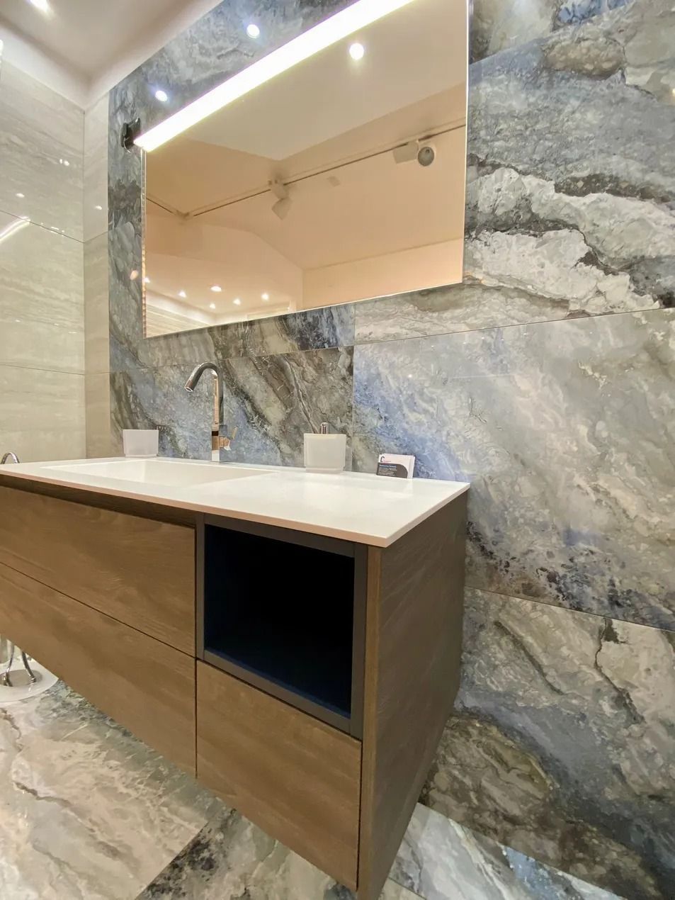 Mobile bagno moderno con mobiletto in legno, piano d'appoggio bianco e ampia parete a specchio con sfondo in piastrelle di pietra.
