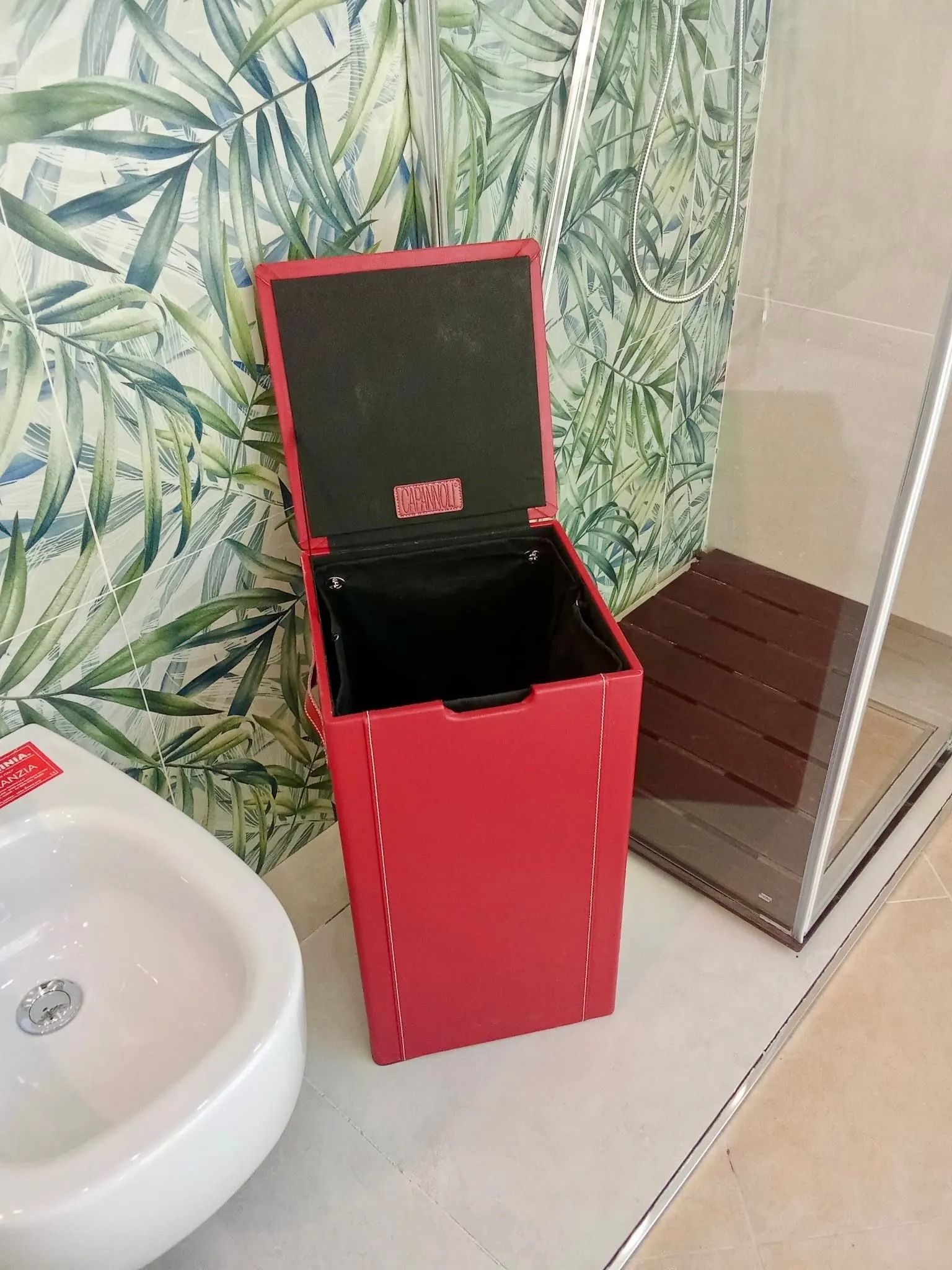 Cestino rosso accanto a un lavandino bianco in un bagno piastrellato con carta da parati a motivi floreali.