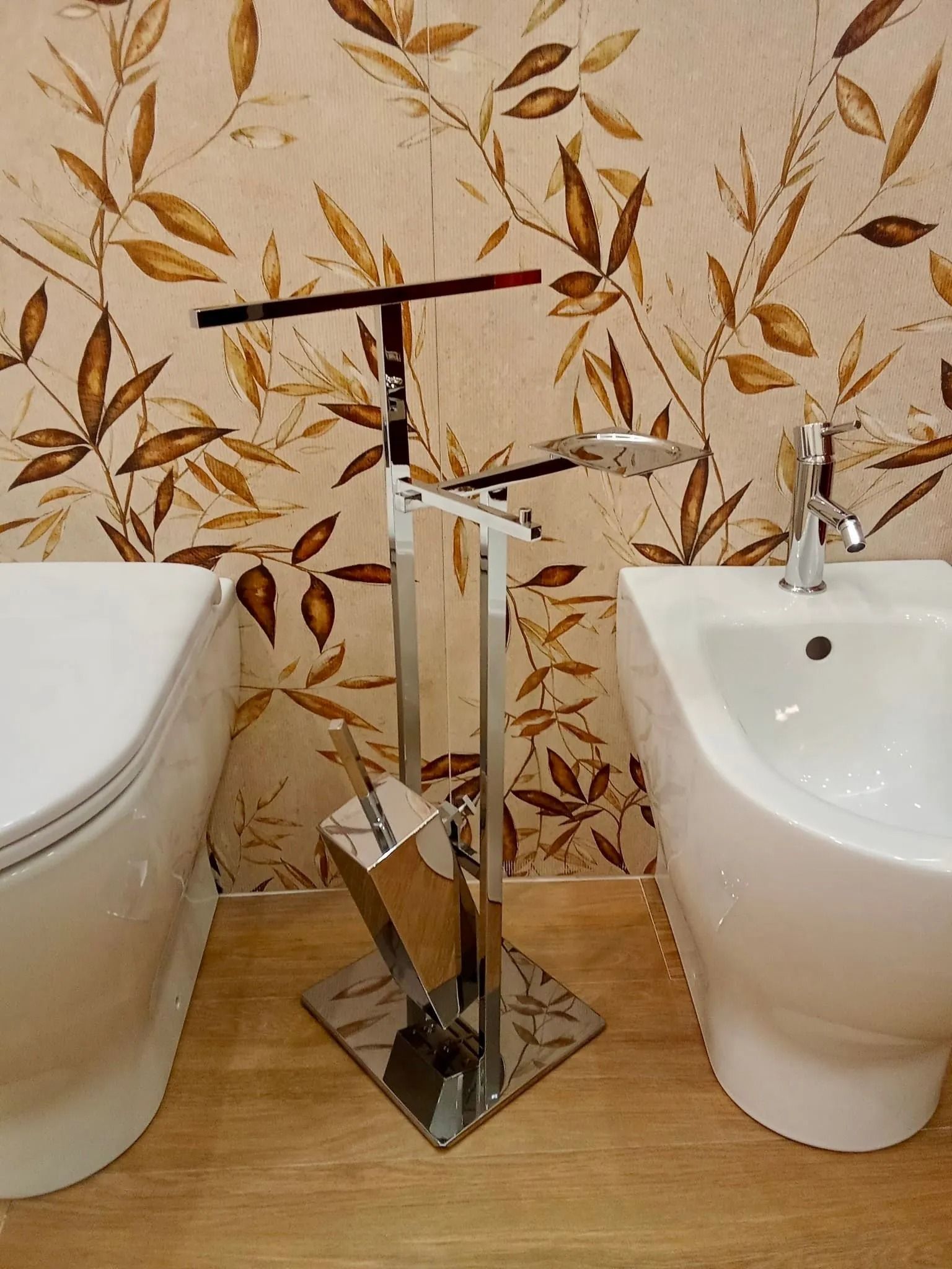 Portascopino cromato tra il WC e il lavandino in un bagno con carta da parati floreale