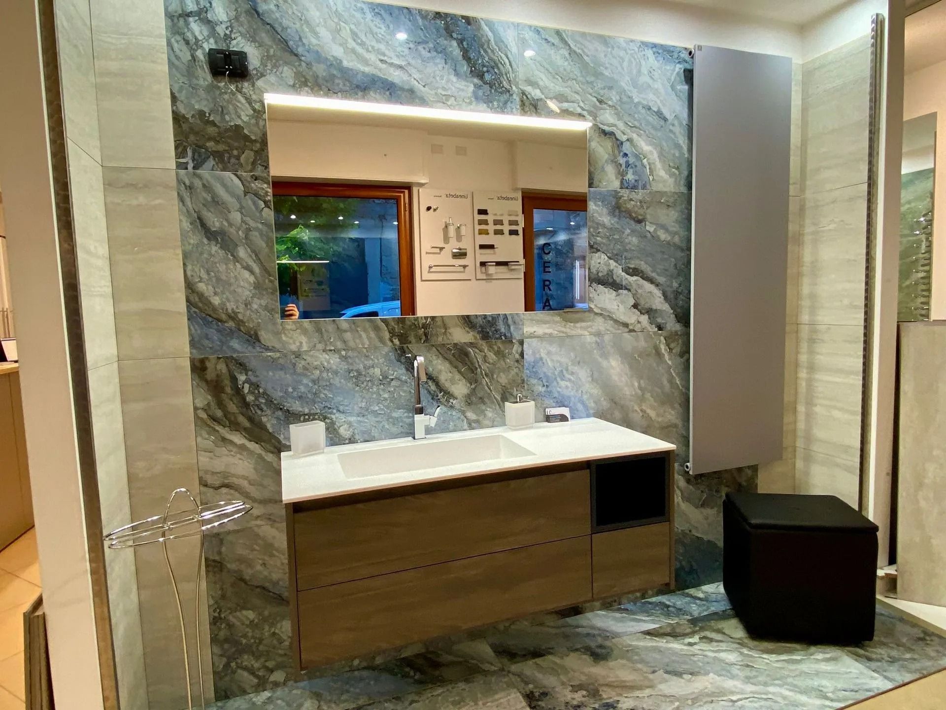Mobile bagno moderno con pareti in marmo, mobiletto in legno, lavabo, specchio e piccolo sgabello nero.