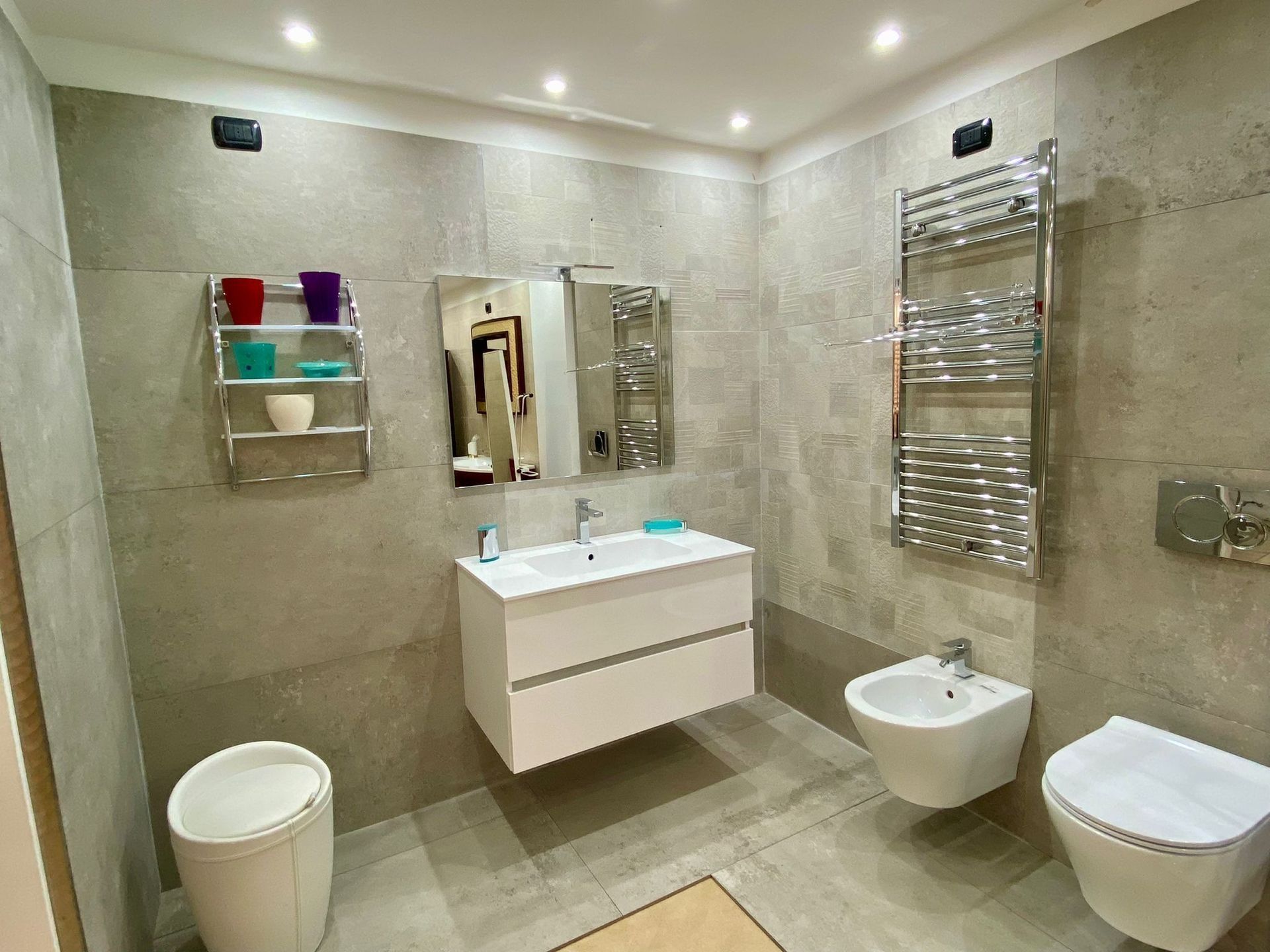 Bagno moderno con piastrelle grigie, mobile lavabo sospeso, specchio a parete, WC, bidet, scaldasalviette e ripiano per la doccia.