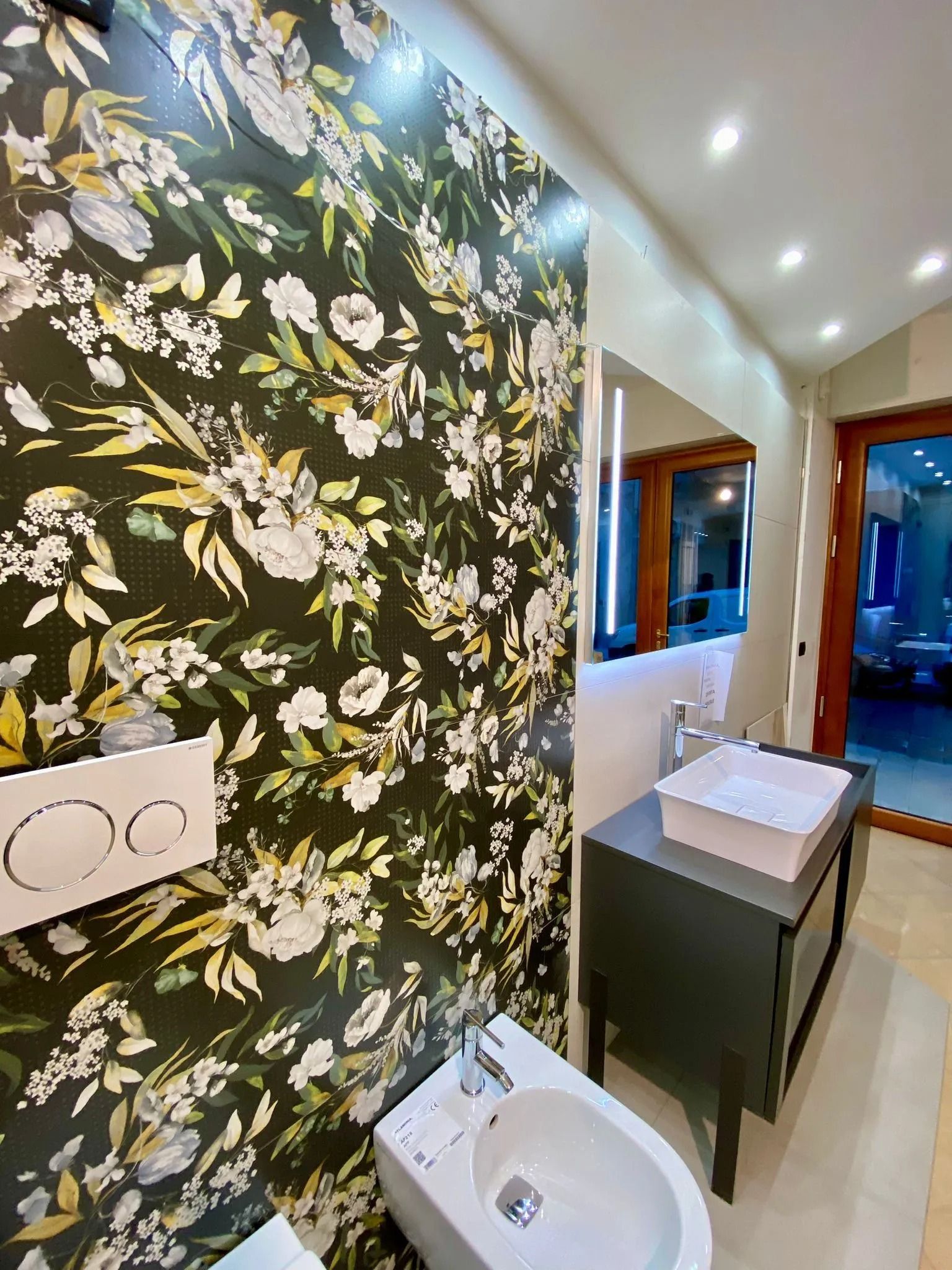 Bagno moderno con carta da parati floreale, WC bianco, lavabo, specchio e vista dalla finestra.