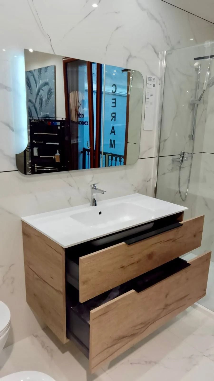 Mobile bagno moderno con lavabo bianco, cassetti in legno e specchio a parete con rivestimento in piastrelle effetto marmo.