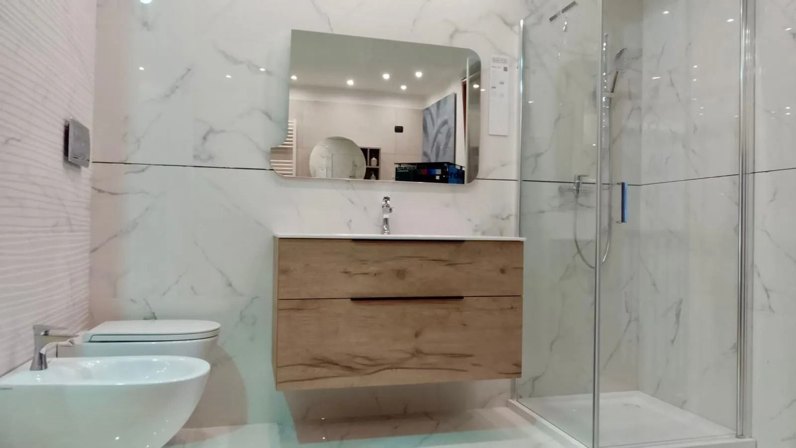 Bagno moderno con pareti in marmo, mobile lavabo sospeso in legno, specchio, doccia in vetro e lavabo e WC a parete.