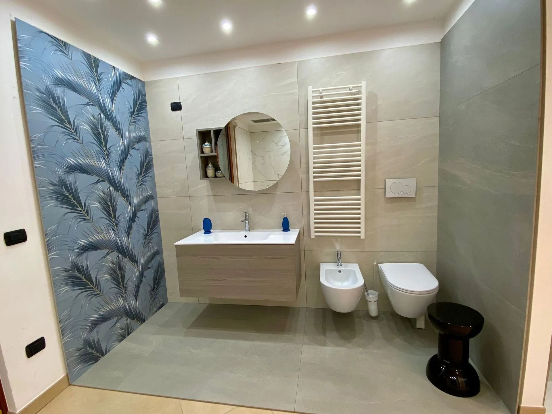 Bagno moderno con murale a tema palme, lavabo sospeso, specchio ovale, bidet, WC e pareti rivestite con piastrelle grigie.