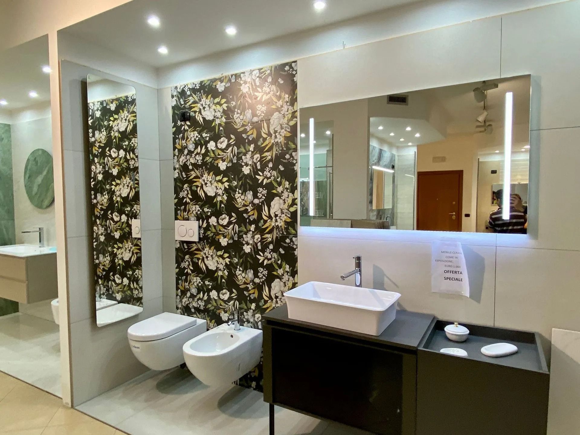 Bagno moderno con parete decorata con motivi floreali, specchio a parete, doppio lavabo e WC e bidet bianchi.