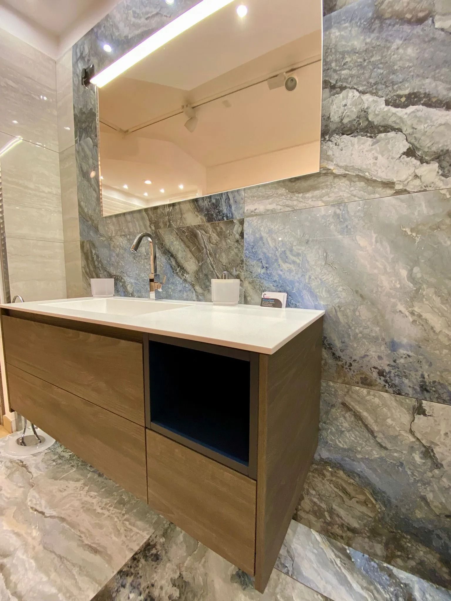 Mobile lavabo in legno con piano bianco e specchio su una parete in marmo grigio in un bagno moderno.