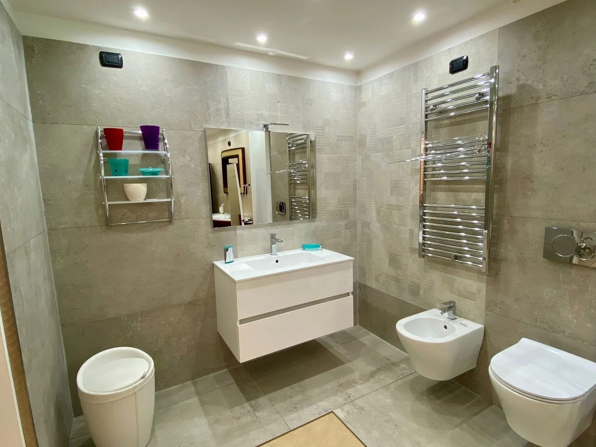 Bagno moderno piastrellato con mobile lavabo sospeso, specchio, WC, bidet e scaldasalviette.