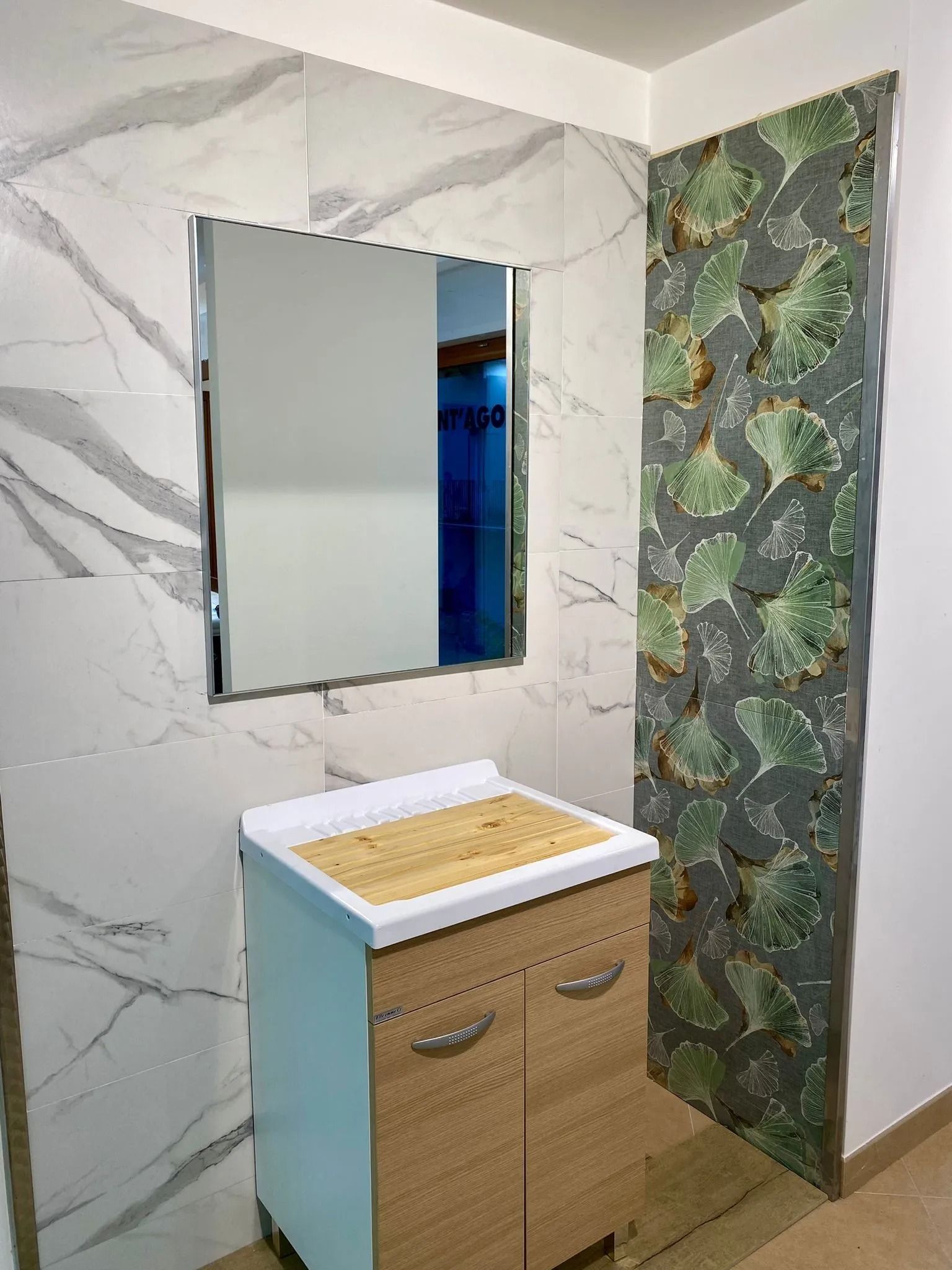 Mobiletto da bagno compatto con specchio, pareti bianche effetto marmo e una parete decorativa con motivo verde.