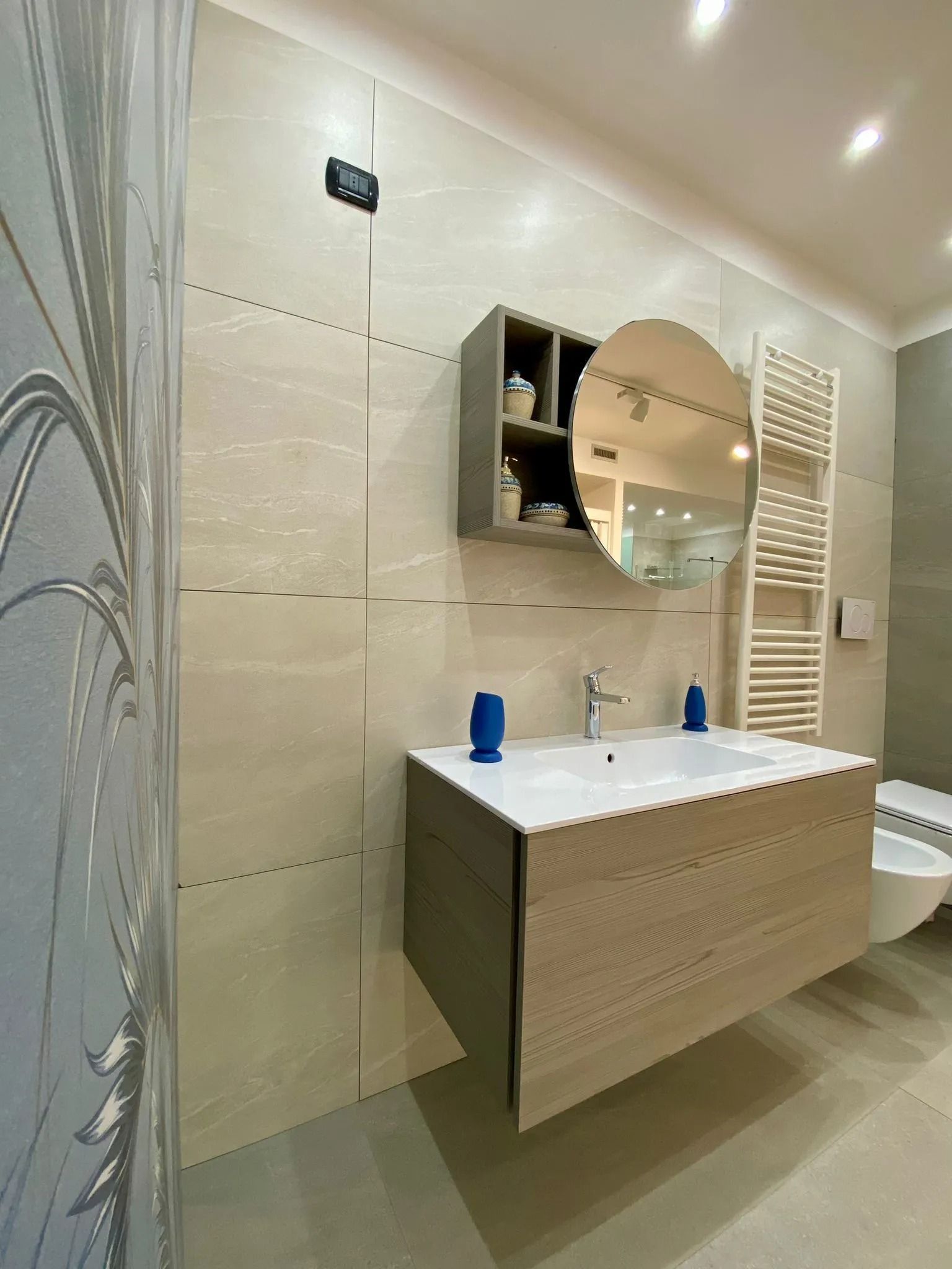 Mobile bagno moderno con specchio rotondo, lavabo bianco e pareti e pavimento rivestiti con piastrelle beige.