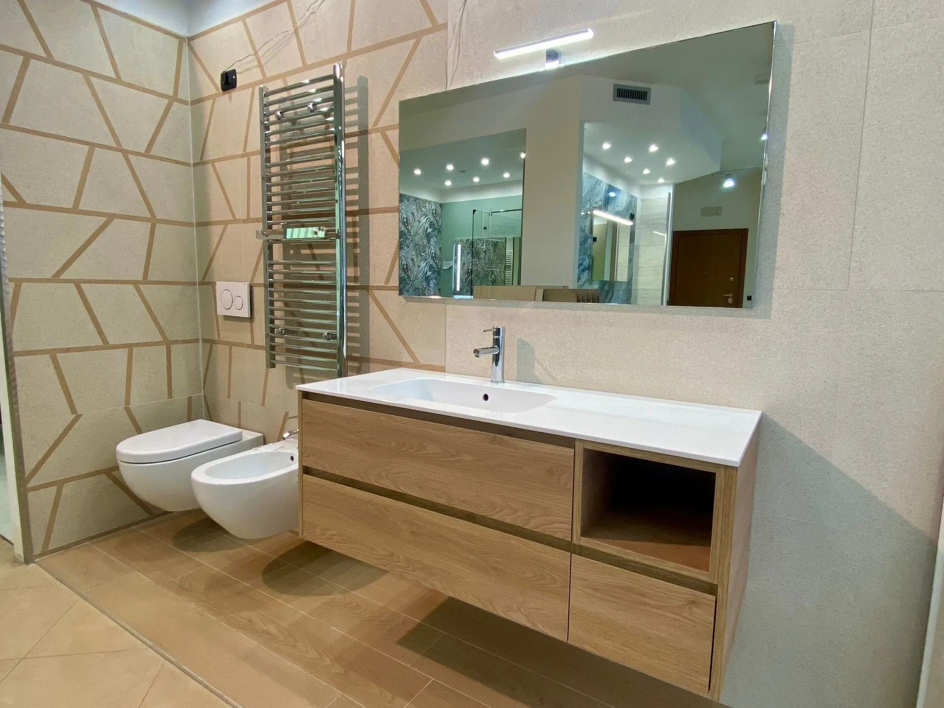 Bagno moderno con WC sospeso, bidet, mobiletto in legno, ampio specchio e pareti rivestite con piastrelle beige.