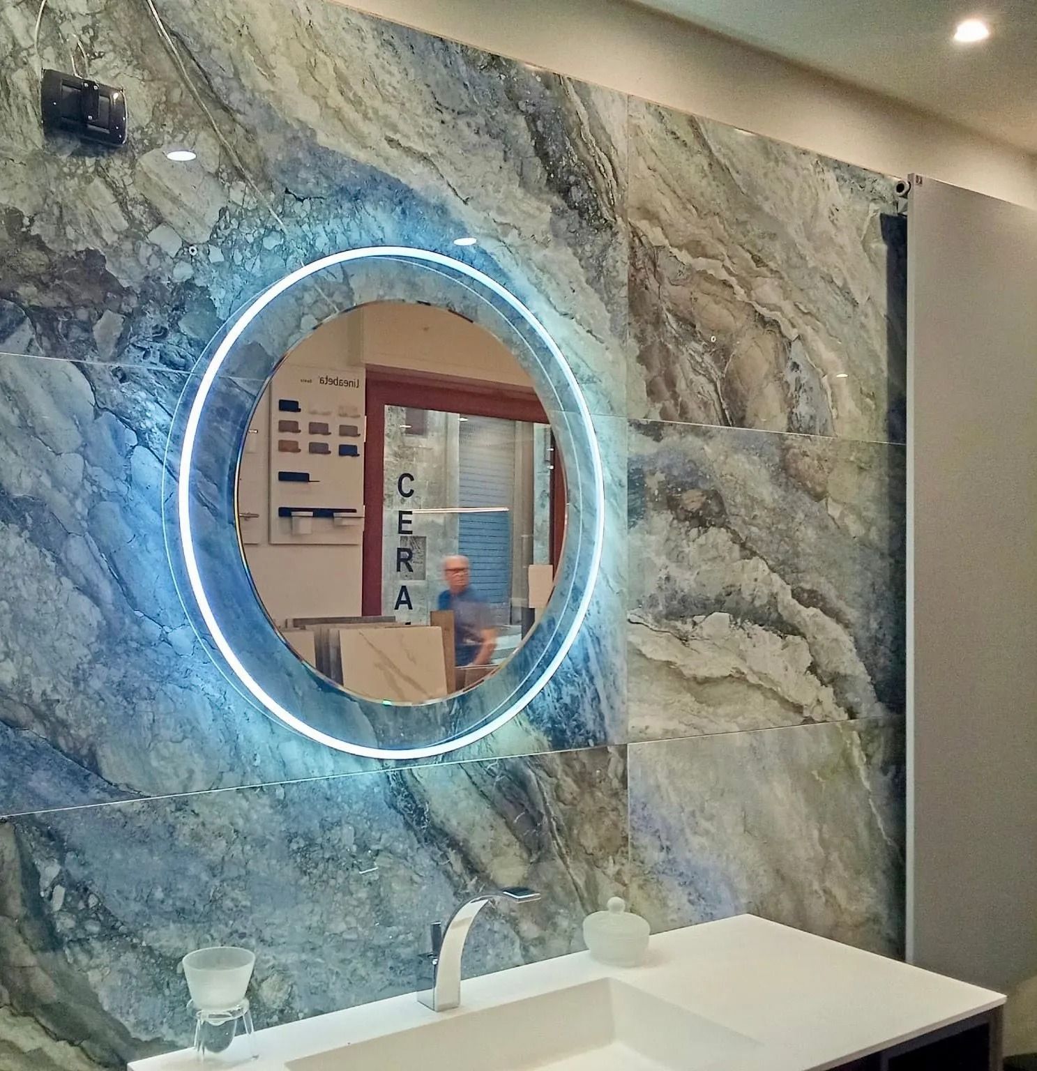 Mobile bagno moderno con parete in marmo, specchio circolare retroilluminato e lavabo bianco.