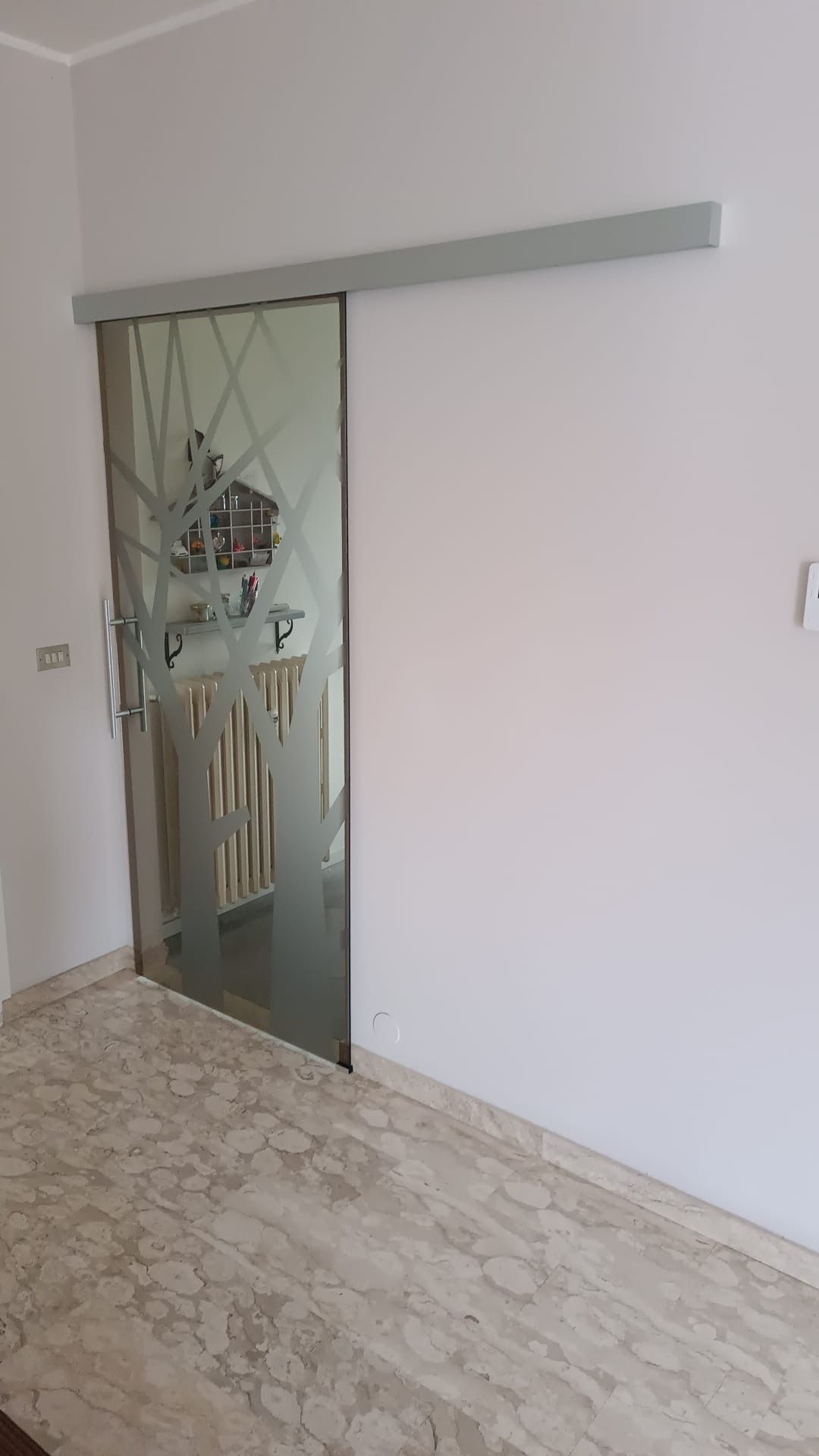 Porta scorrevole in vetro con motivo inciso, montata su binario. Pareti bianche e pavimento in marmo.