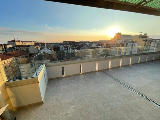 Terrazza panoramica con vista sulla città al tramonto. Pareti beige, ringhiere in vetro.