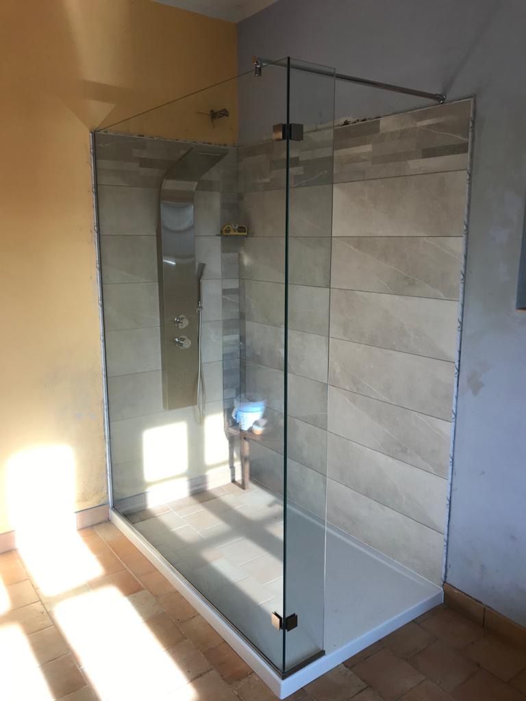 Box doccia in vetro con pareti piastrellate grigio chiaro e base bianca, in una stanza illuminata dalla luce del sole.