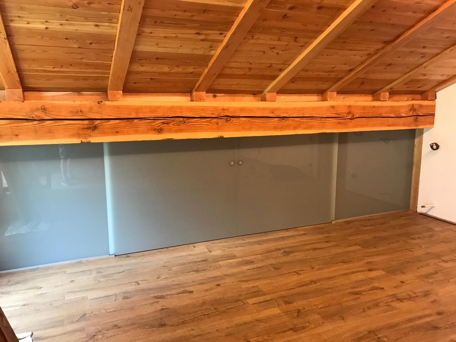 Camera con pavimento in legno, travi a vista sul soffitto e parete con pannelli in vetro smerigliato grigio.