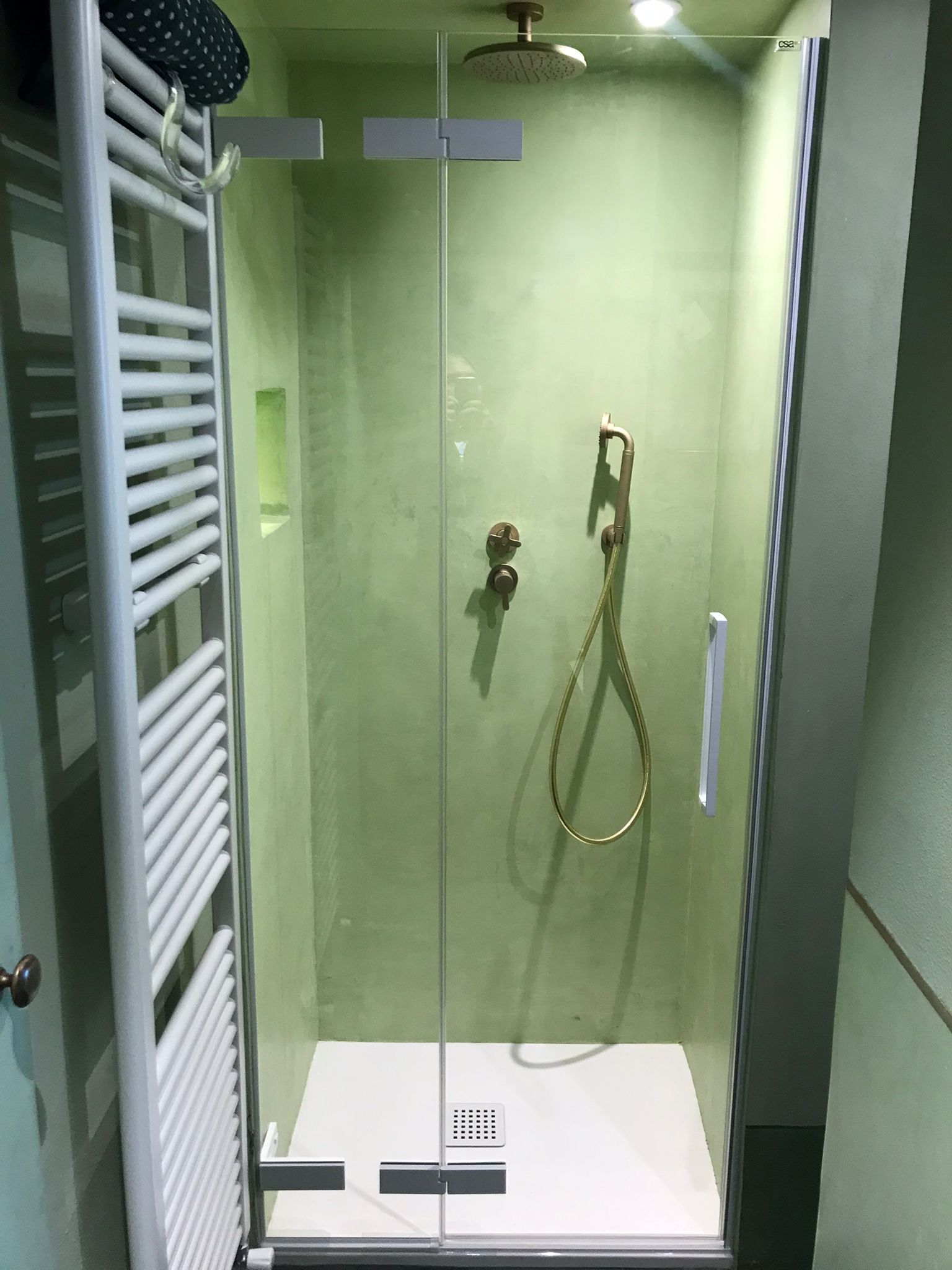 Box doccia con porte in vetro, accessori dorati e base bianca contro una parete verde