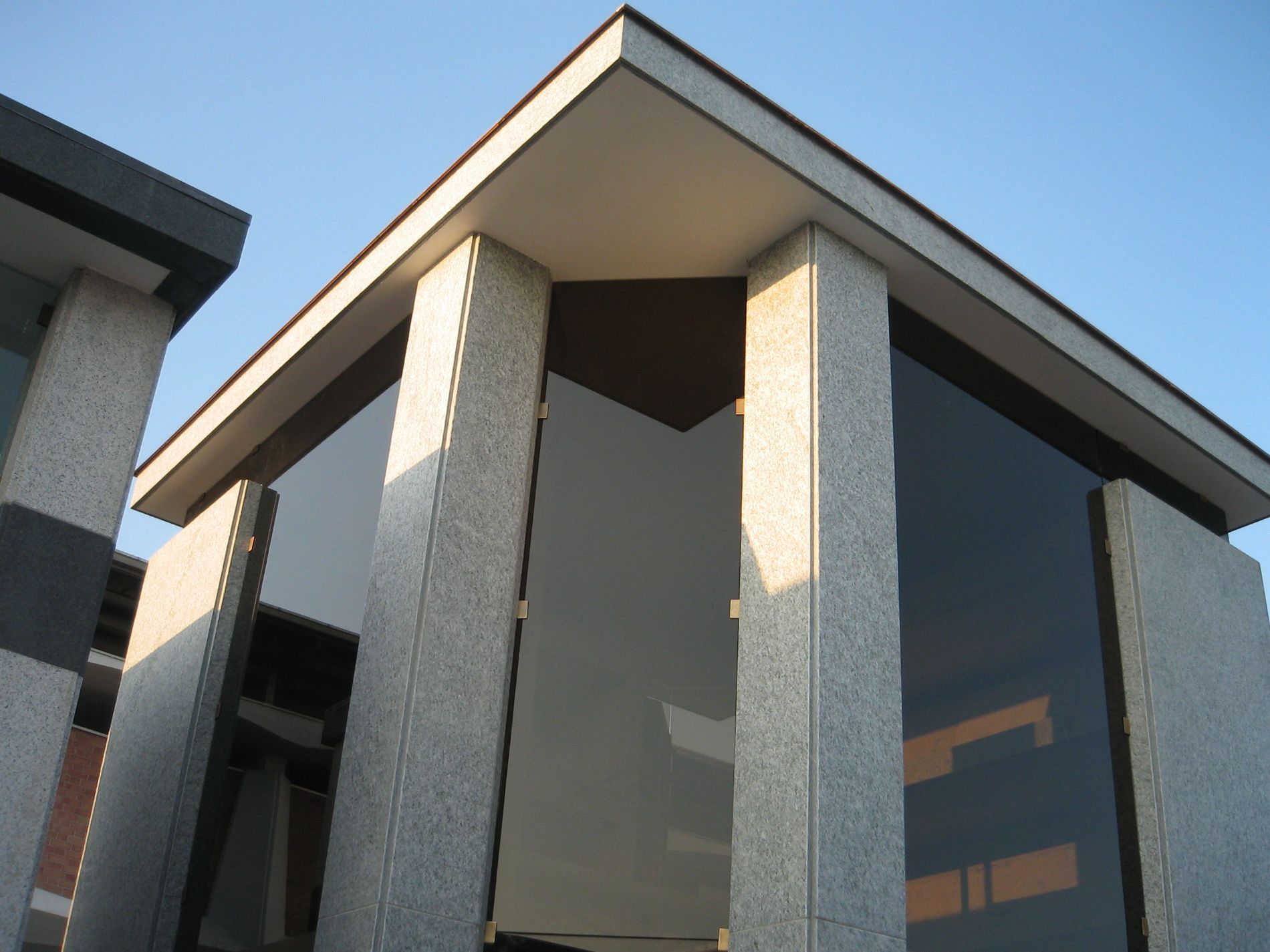 Angolo di un edificio moderno con grandi vetrate, fiancheggiato da pilastri in pietra, sotto un tetto bianco e marrone.