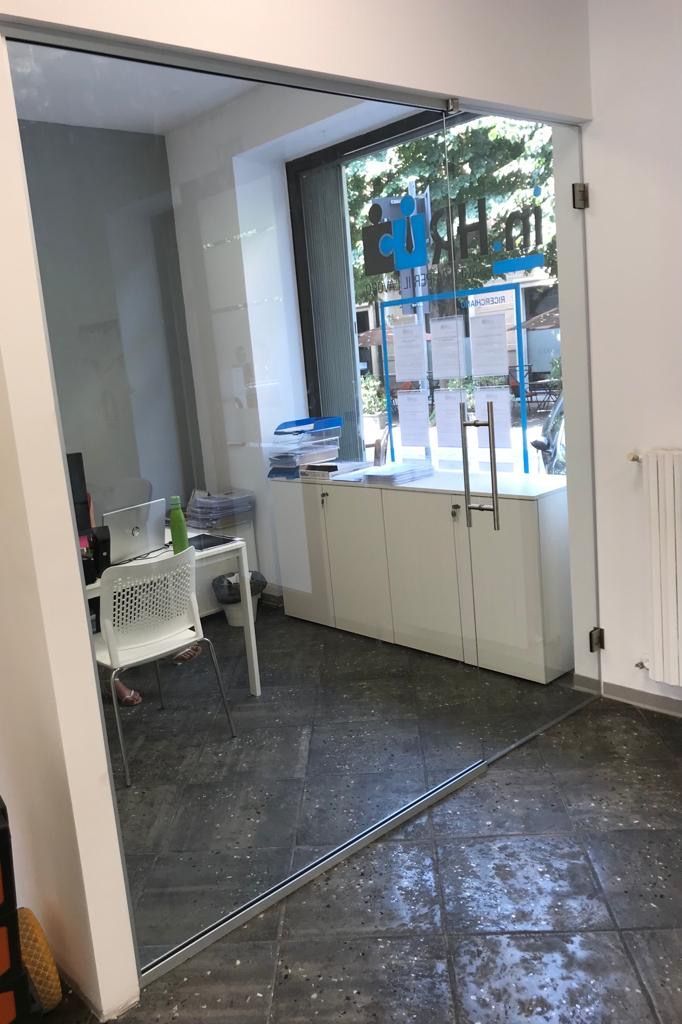 Porta a vetri che conduce a un ufficio; sono visibili armadietti bianchi, scrivania, sedia e una grande finestra